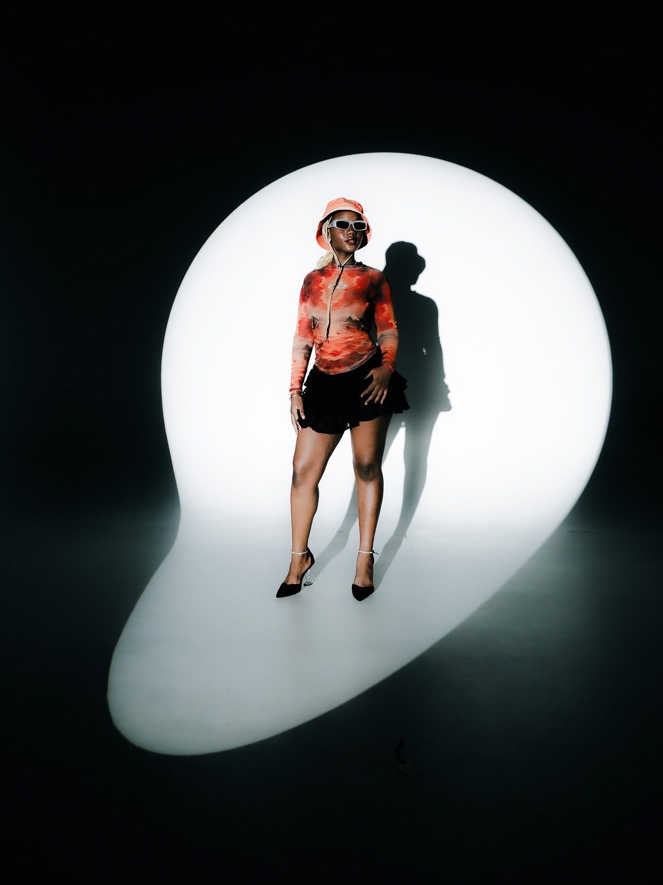 A woman in red top, black shorts, and heels poses dramatically in front of a glowing white light orb on dark background.