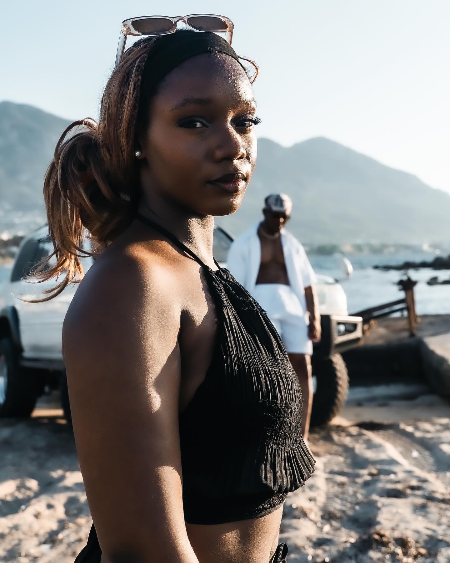 Young woman in black halter top with sunglasses on head stands on beach, mountains and truck visible behind her.