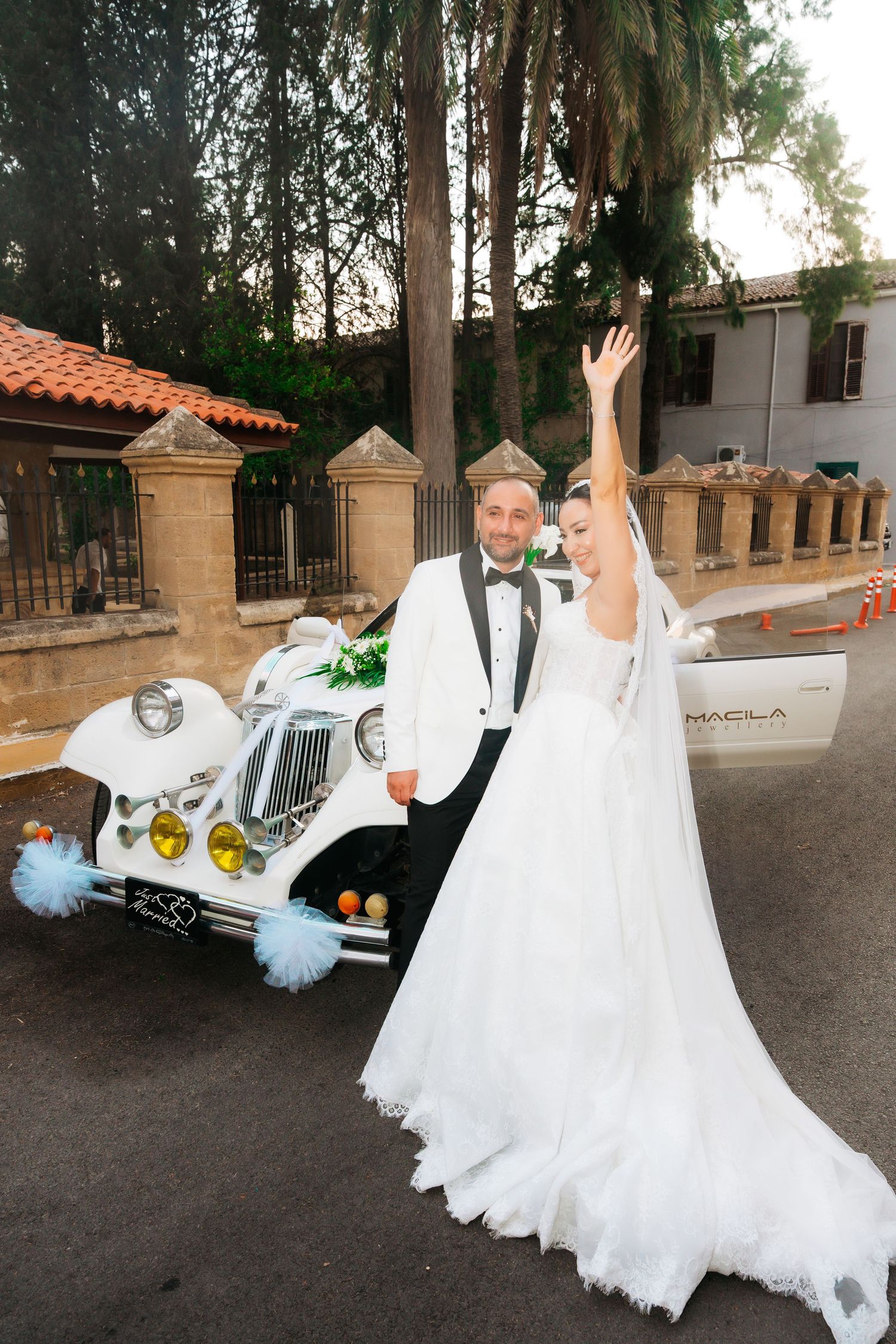 Bride in white gown raises arm joyfully beside groom in tuxedo next to decorated vintage white wedding car.
