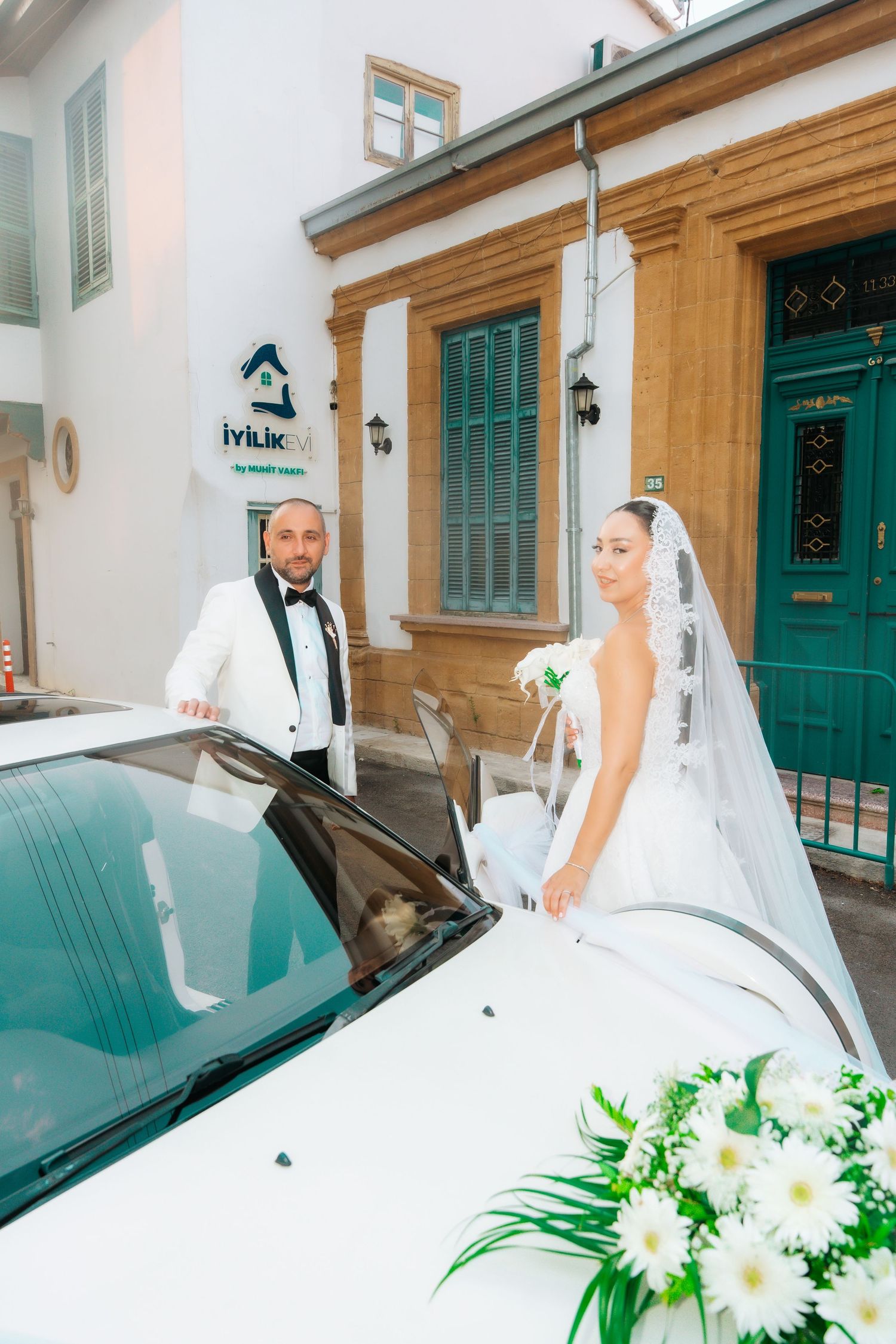 Bride and groom in formal attire standing beside a white flower-decorated wedding car outside a building.