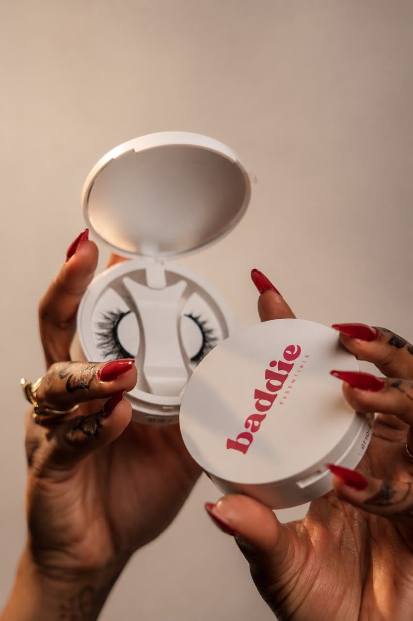 Hands with red nails hold open a white Baddie brand compact case containing false eyelashes.
