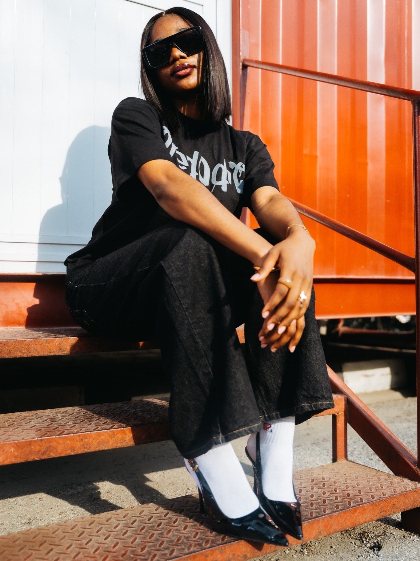 A woman in a black graphic tee, wide-leg jeans, white socks, and black heels sits on metal steps outdoors.