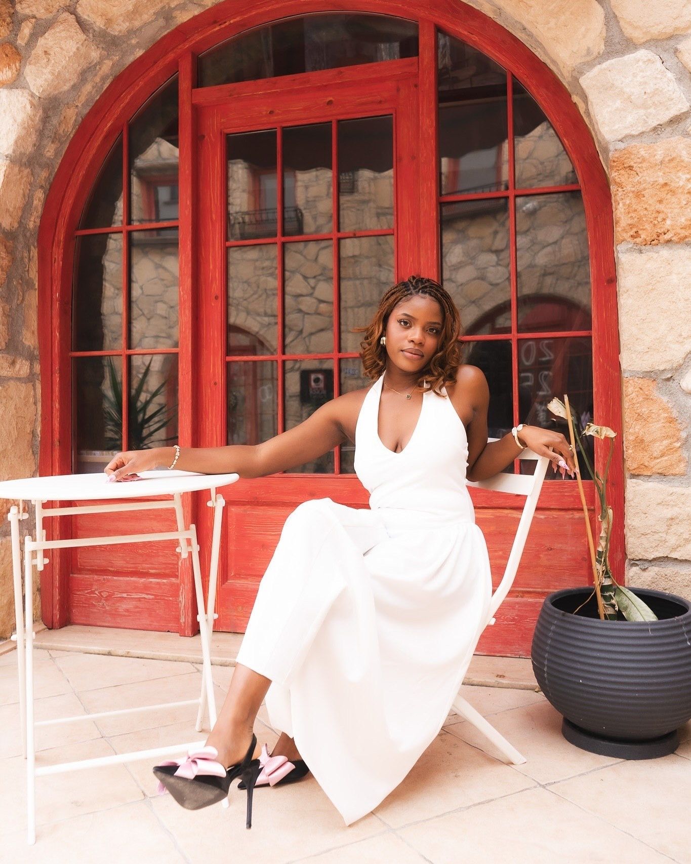 Woman in white halter dress seated at outdoor cafe table beside red arched window and black planter.