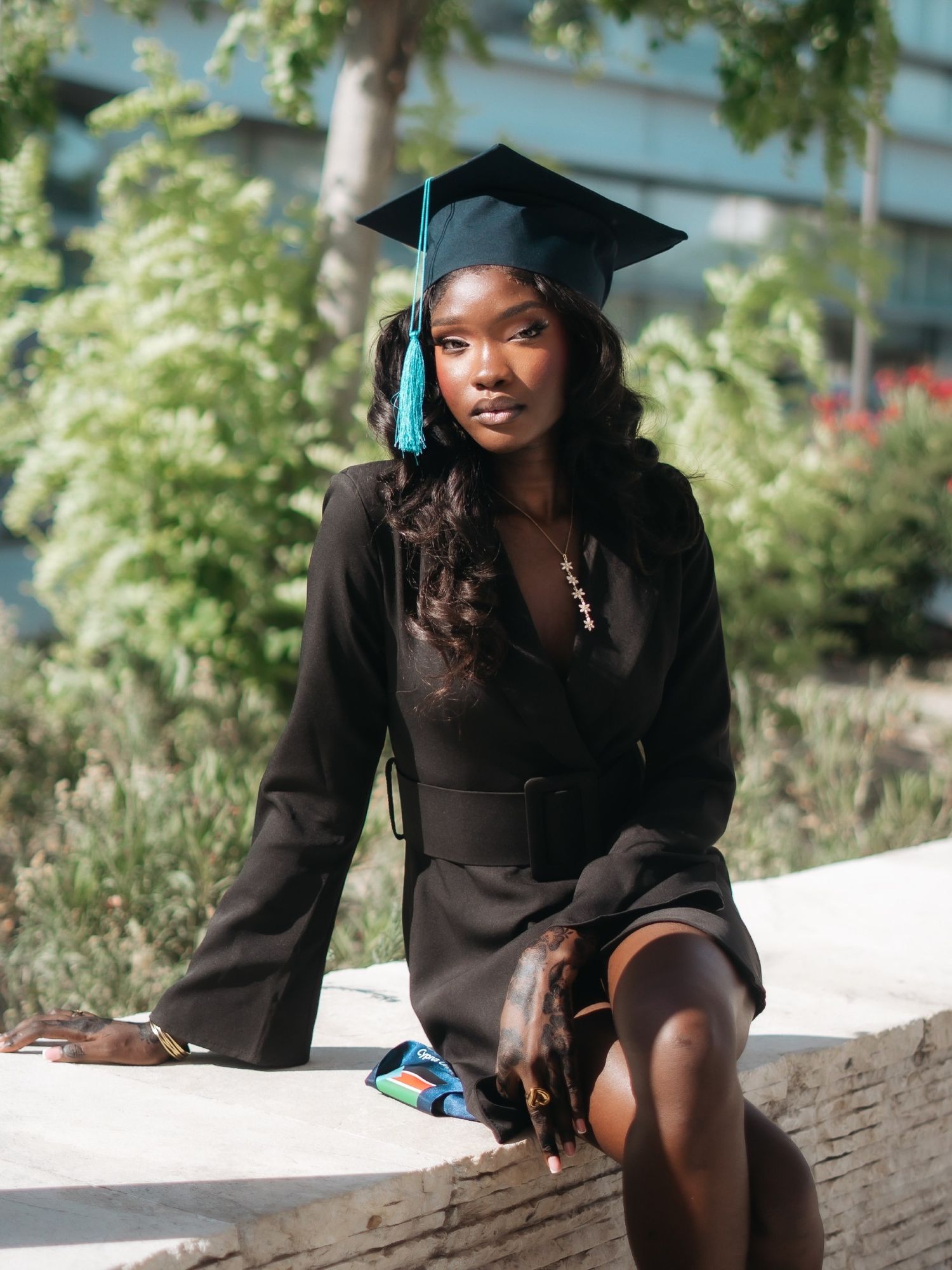 Graduate in black blazer dress and teal-tasseled cap sits outdoors in sunlight.