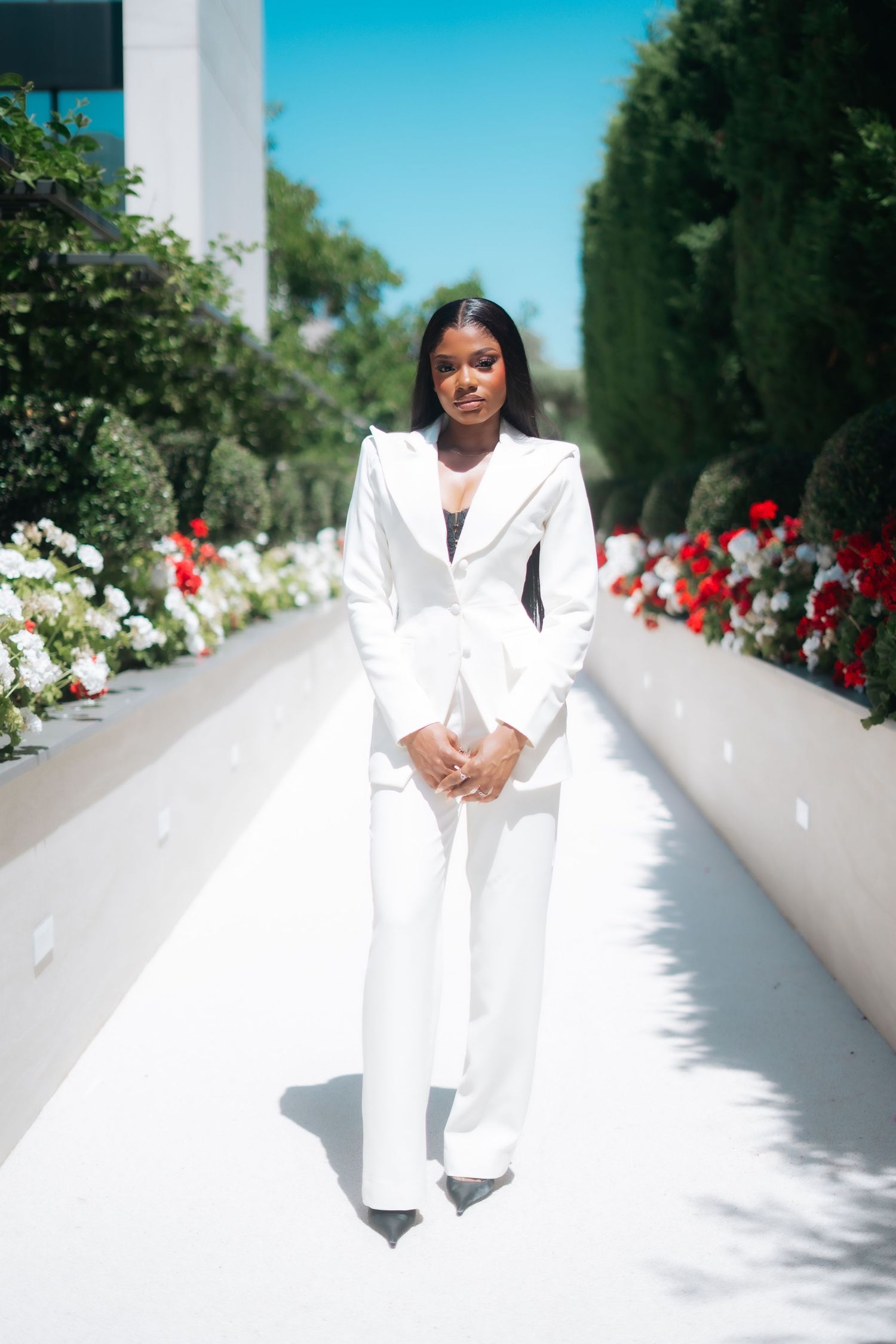 Elegant Black woman in white pantsuit stands in sunny outdoor garden pathway with flowers.