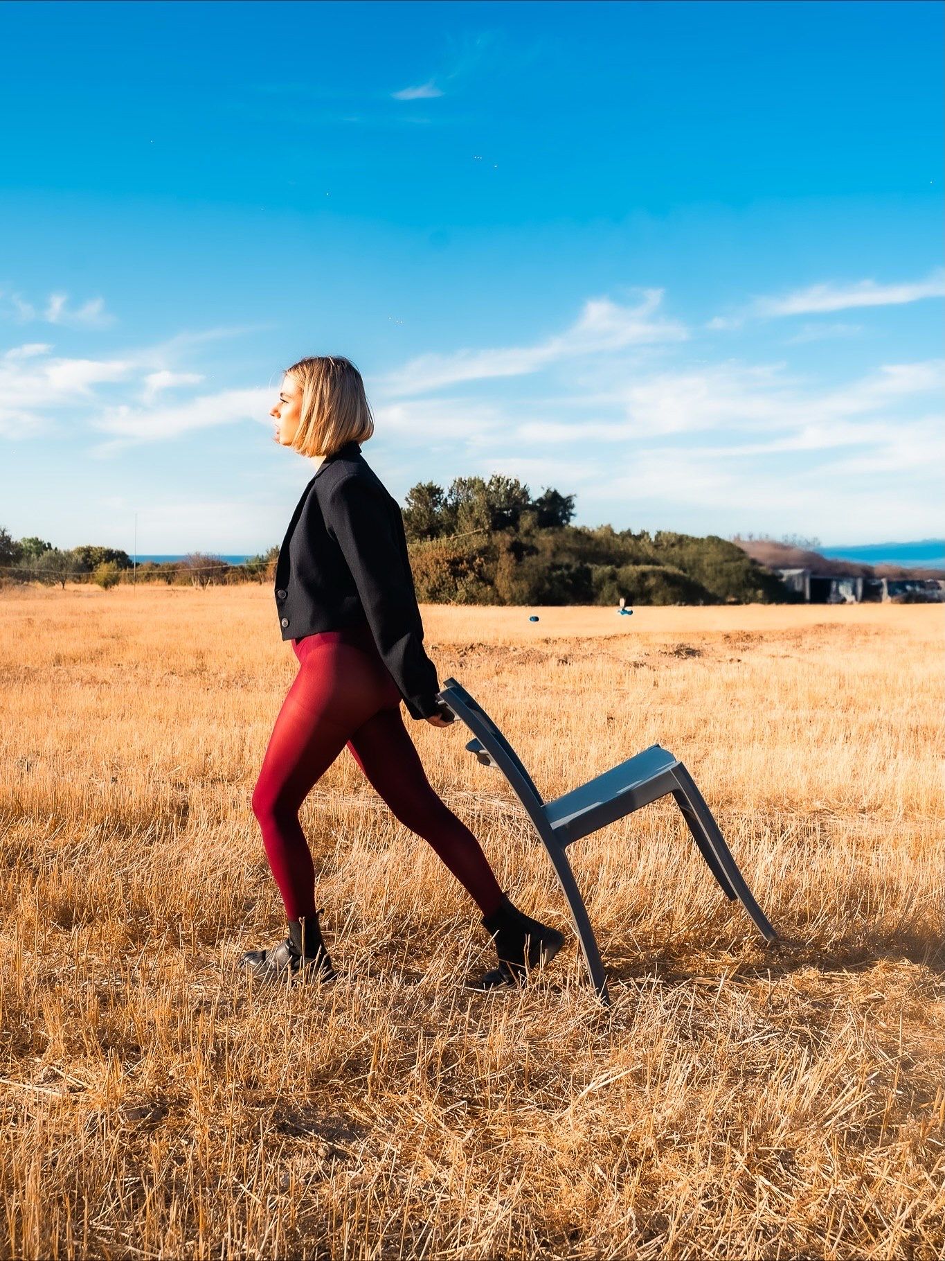 Blonde woman in red leggings and black jacket dragging a chair through a golden field under a blue sky.