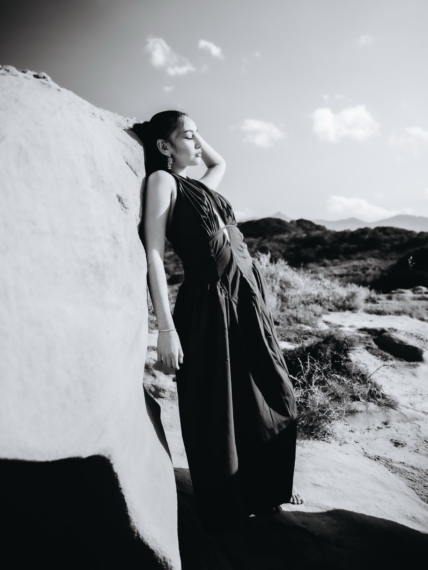 Black and white photo of a woman in a dark dress leaning against a large rock in a desert landscape.