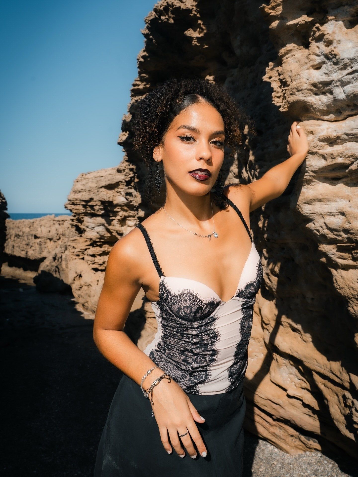 Woman in black lace corset dress poses against rocky coastal cliffs under a clear blue sky.