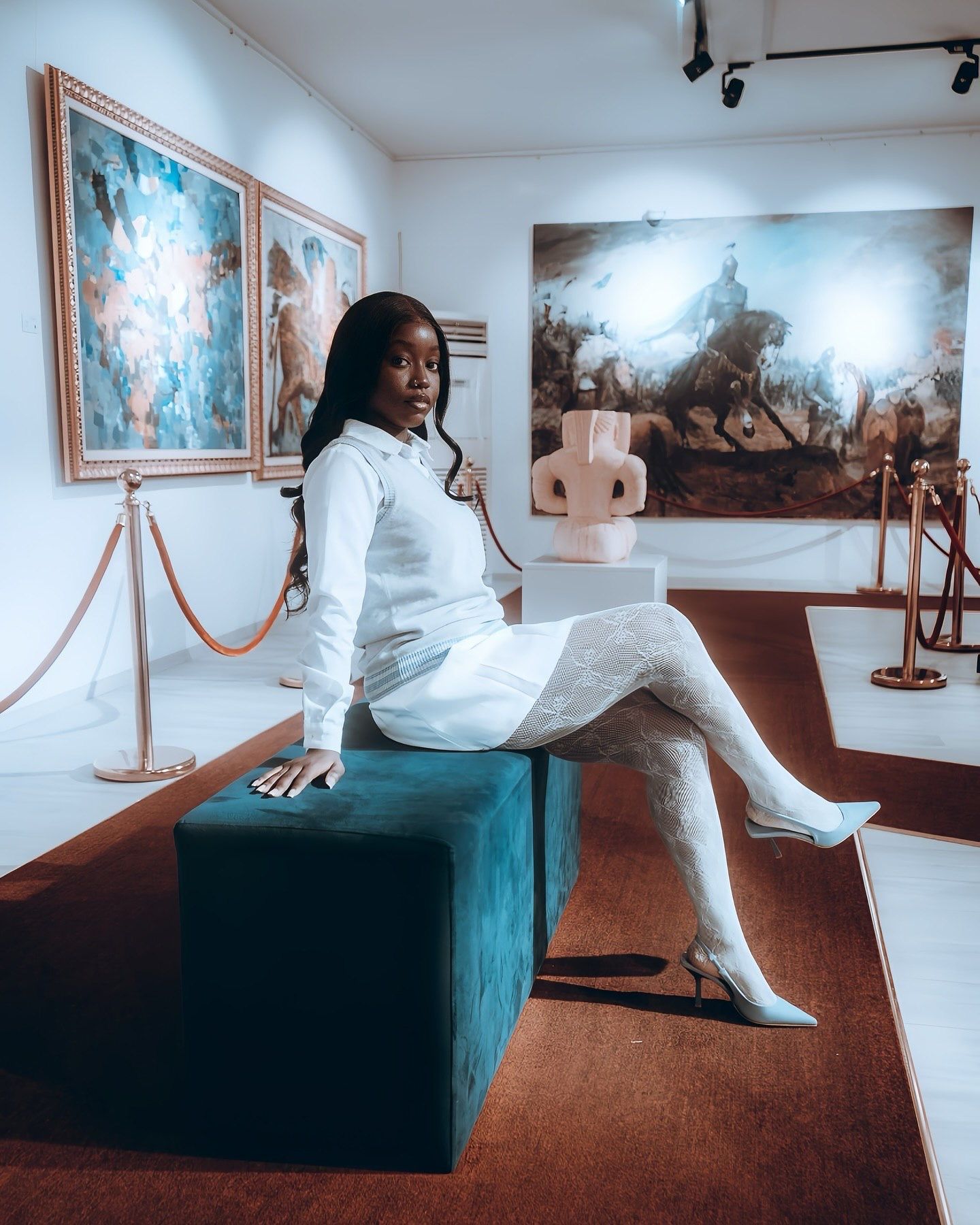 Woman in white outfit poses on teal velvet bench in art gallery surrounded by paintings and sculptures.