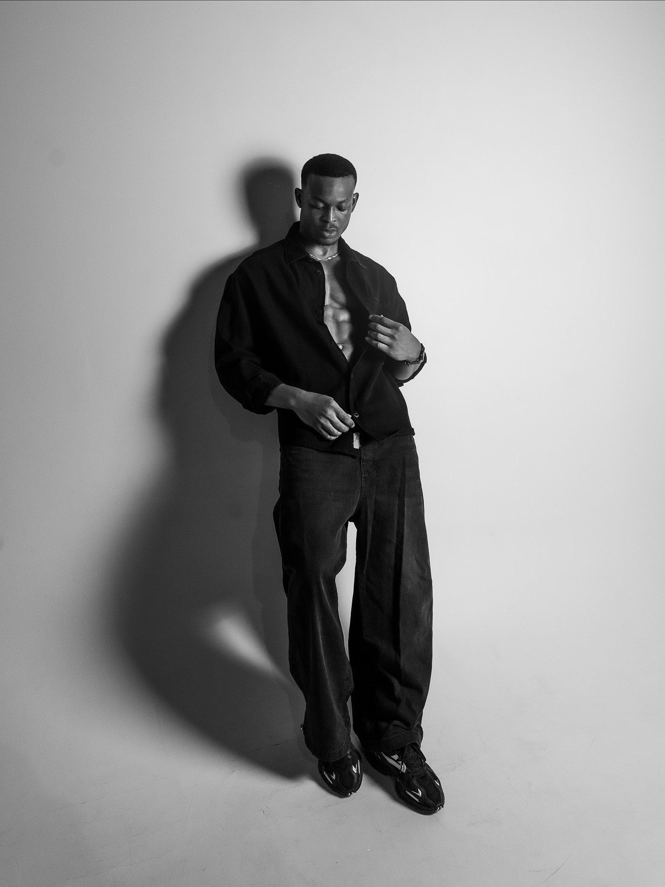 Black and white photo of a man in all-black outfit with open shirt, posing confidently in a studio setting.