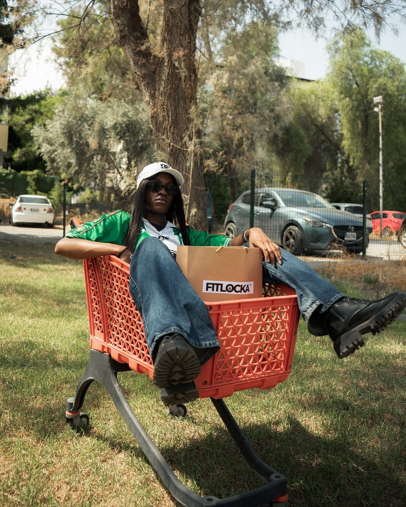 Person relaxing in a red shopping cart outdoors holding a Fitlock box, wearing green shirt, jeans, and black boots.