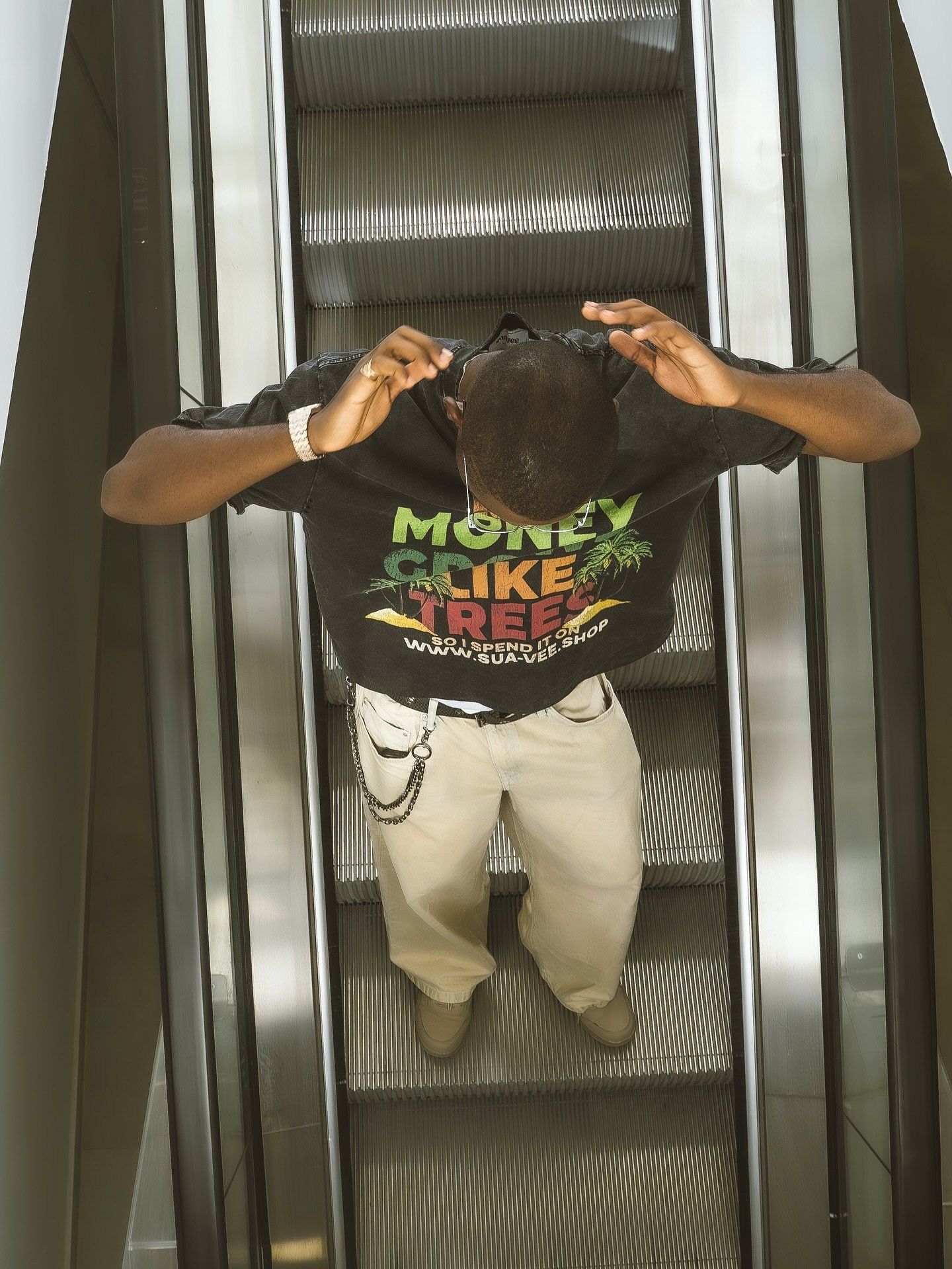 Man on escalator wearing Money Grows on Trees graphic tee, khaki pants, and chain belt, viewed from above.