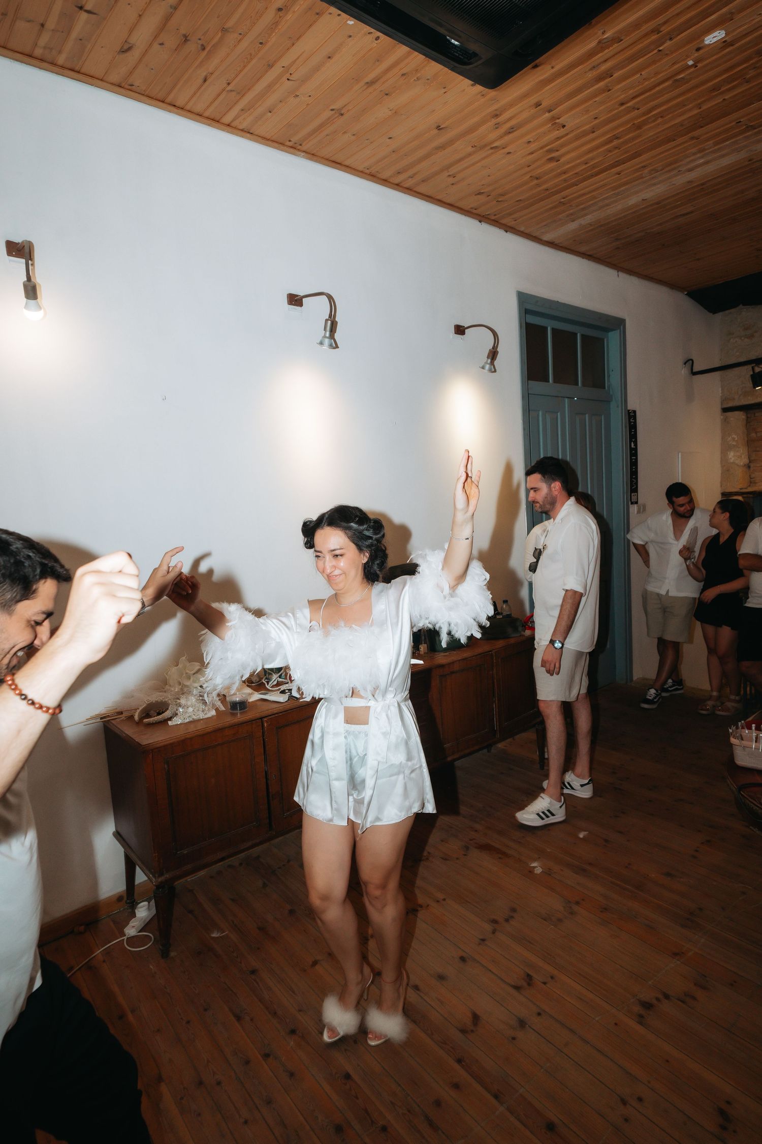 Woman in white feathered outfit dancing joyfully at an indoor party with wooden floors and white walls.
