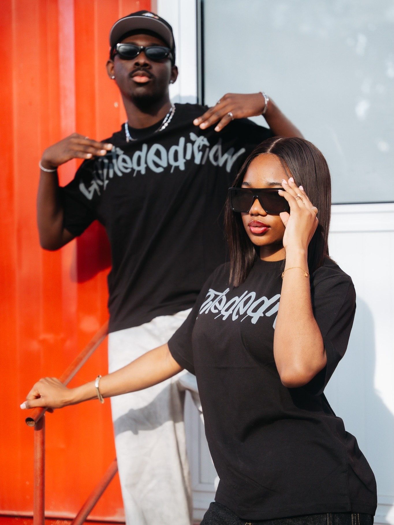 Two stylish people wearing matching black graphic tees and sunglasses posing against a vibrant orange wall.