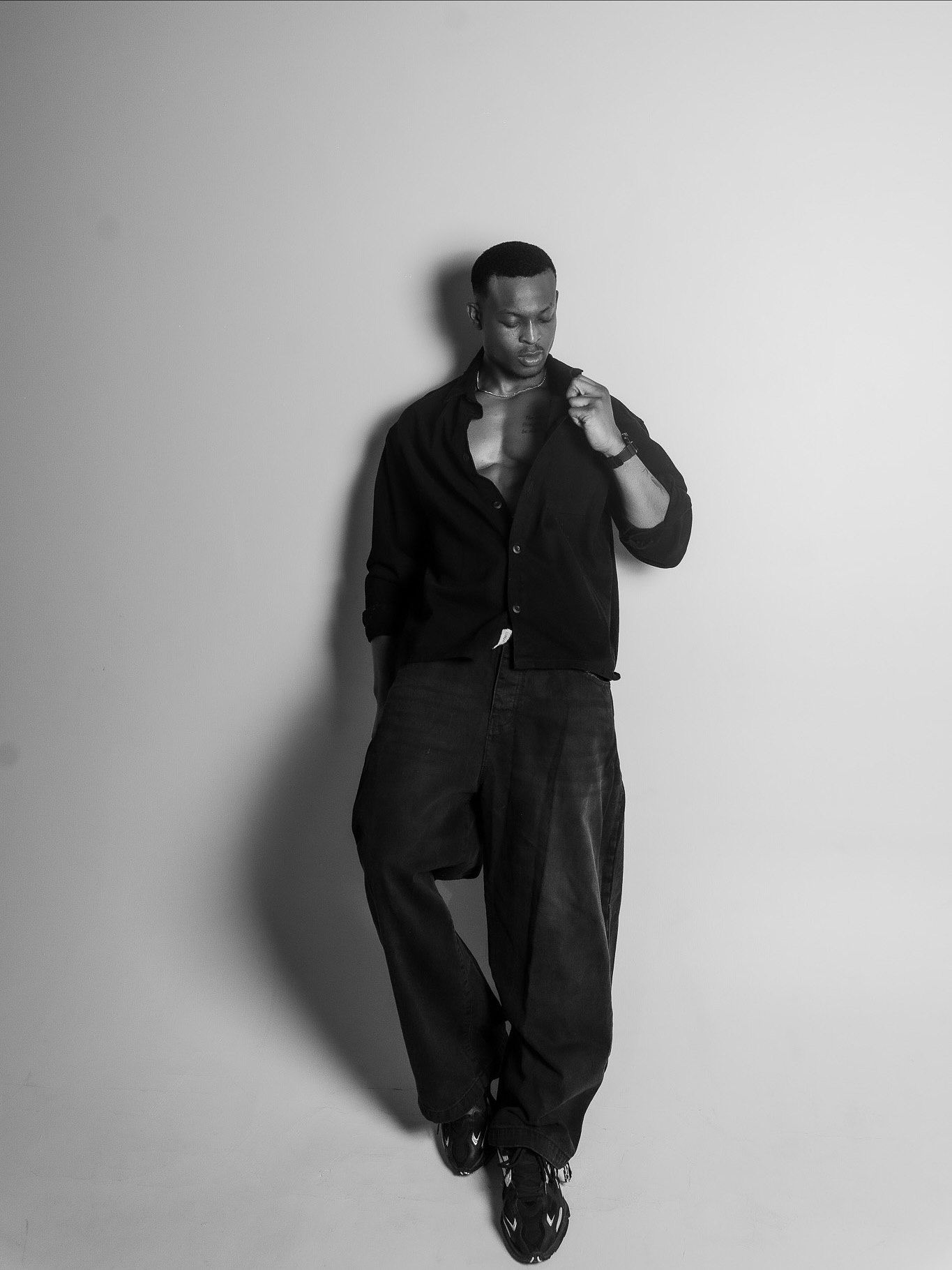 Black and white photo of a stylish man in dark cardigan and wide-leg jeans posing confidently against a light wall.