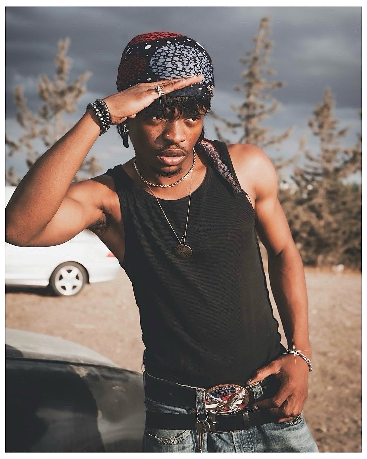 Young man in black tank top, red bandana, and cowboy belt buckle poses outdoors near cars and trees.
