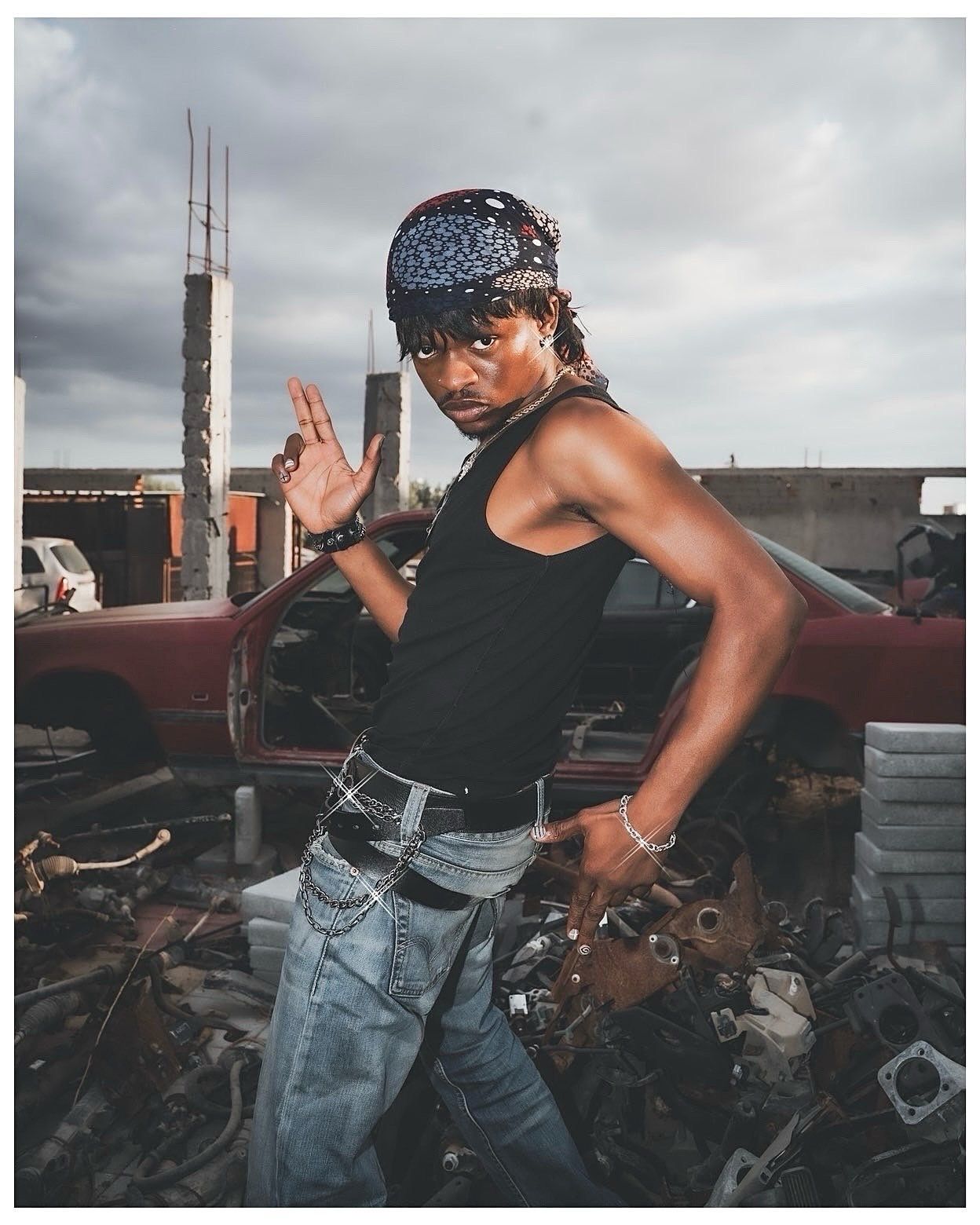 A muscular man in a black tank top and jeans poses confidently in a junkyard with a bandana and chain belt.