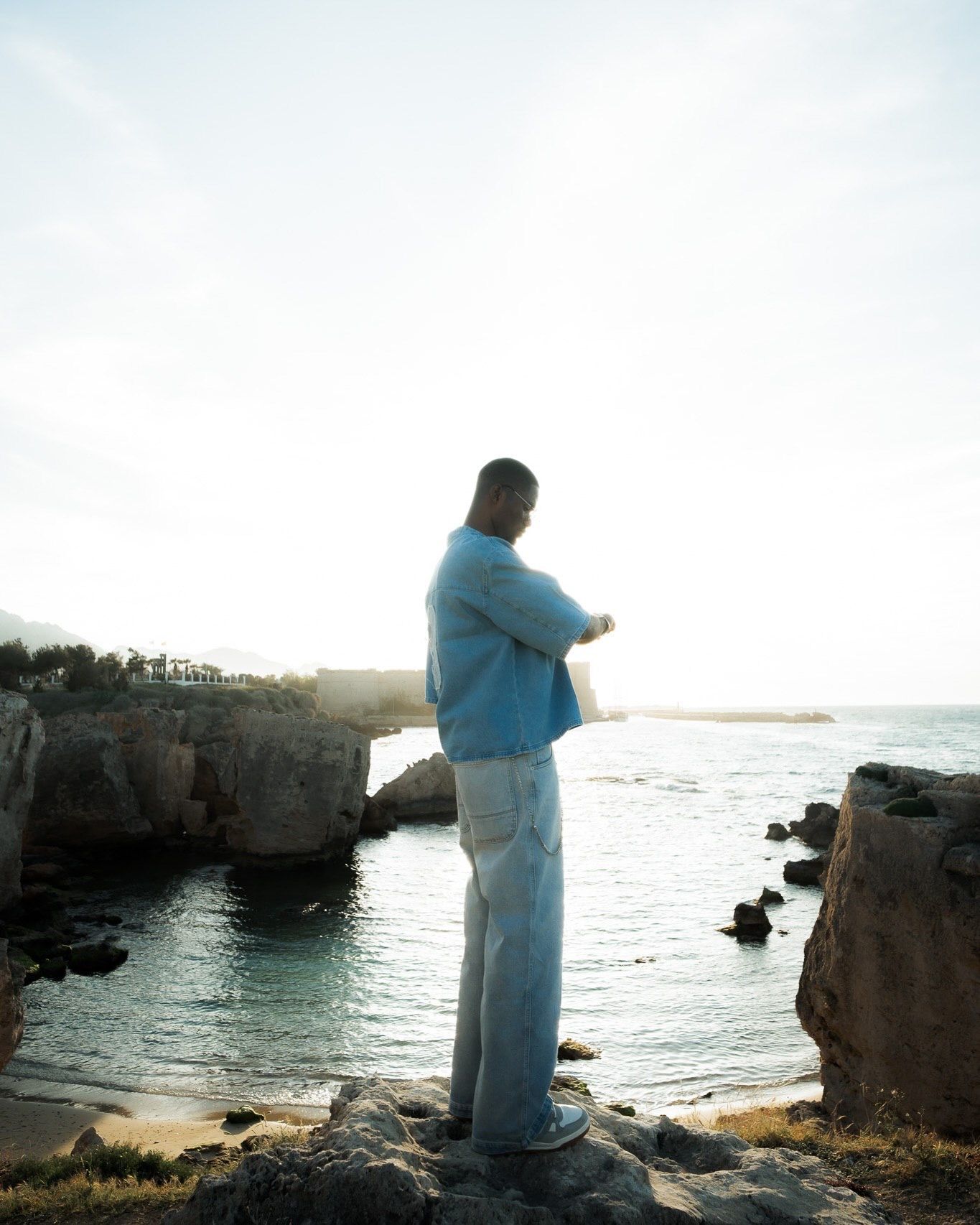 Person in blue sweater and wide-leg jeans standing on rocky coastal shoreline overlooking calm ocean waters.