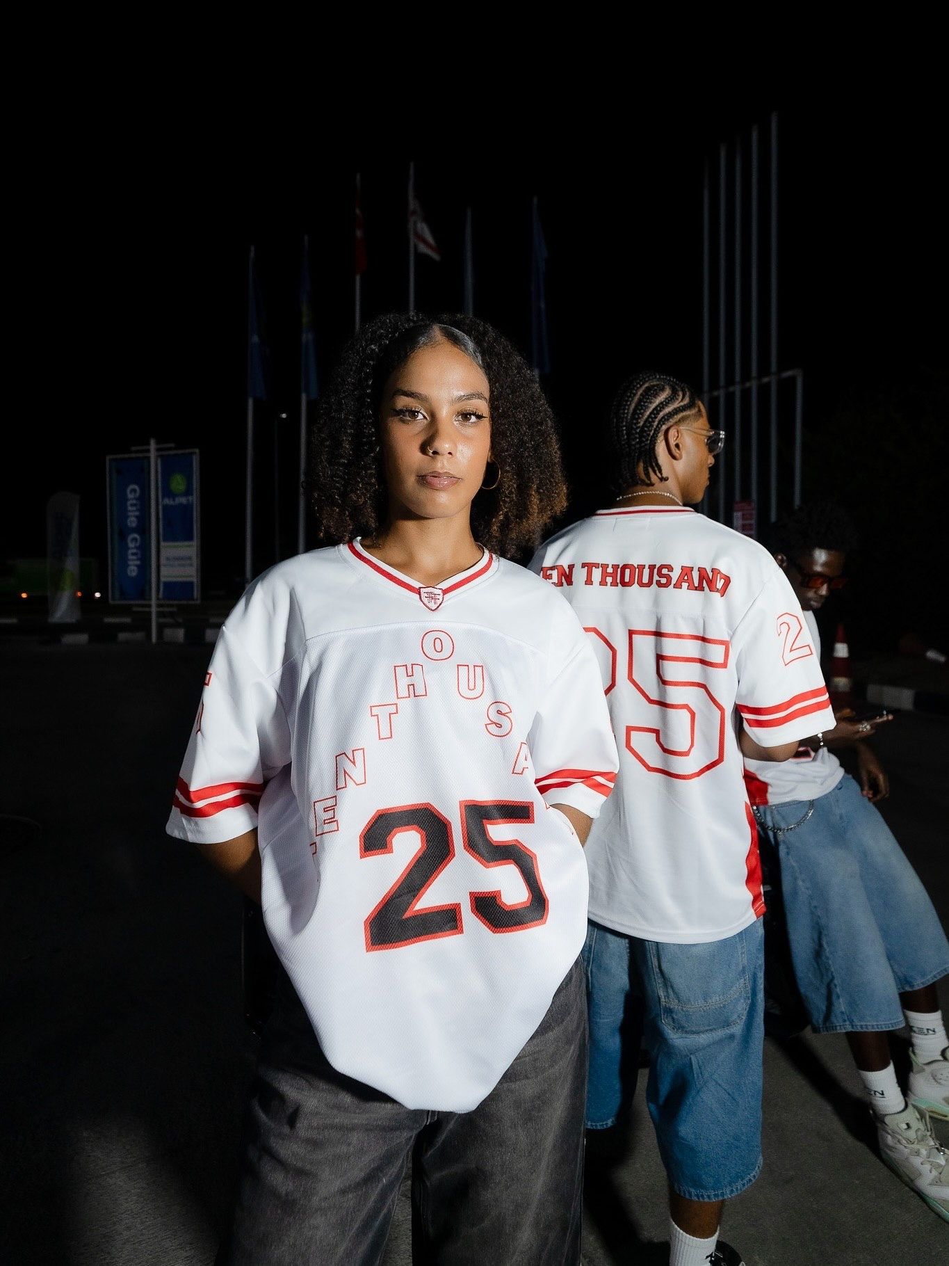Two people wearing white number 25 sports jerseys with red trim outdoors at night.