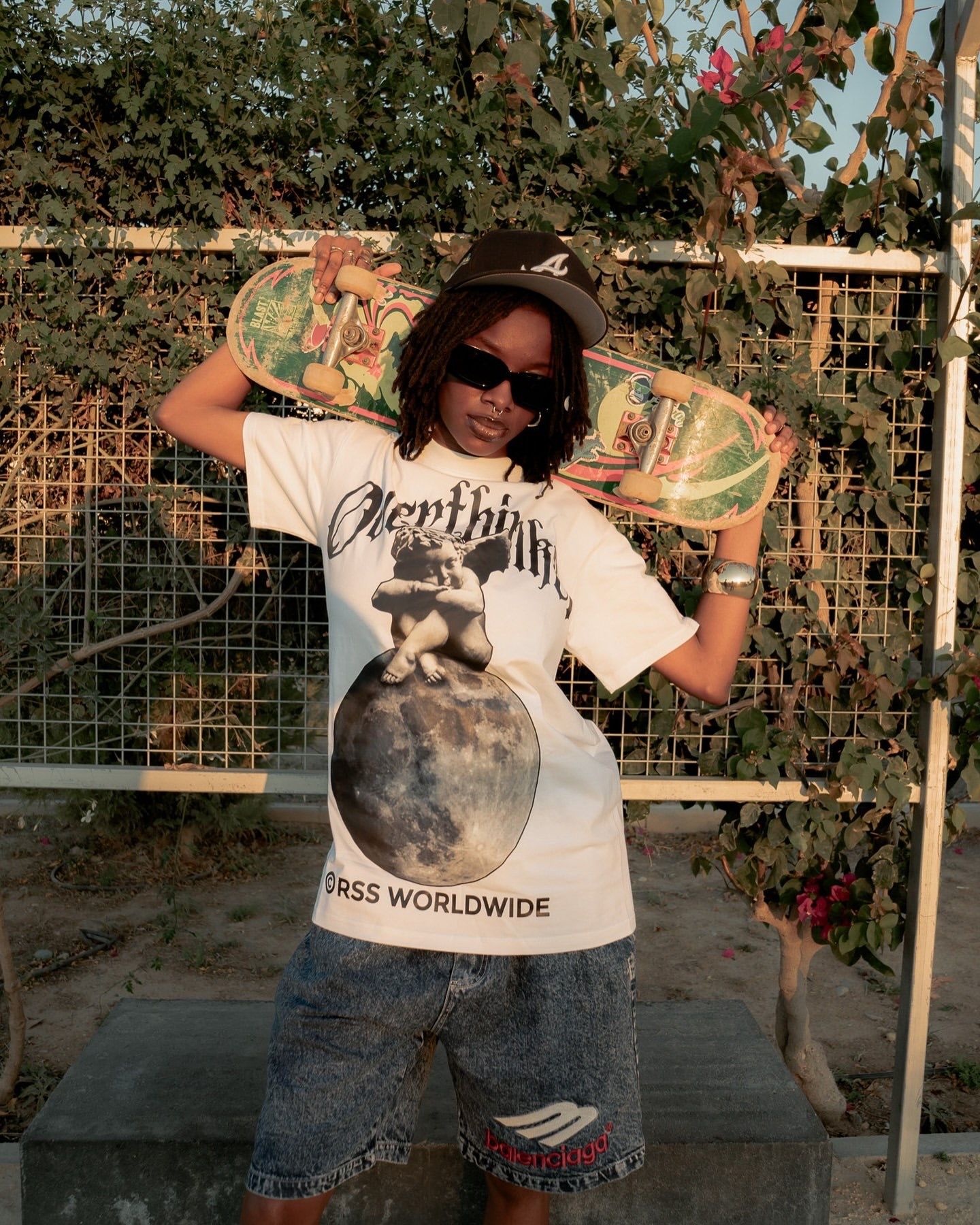 Young woman posing with a skateboard wearing a graphic tee and denim shorts outdoors.