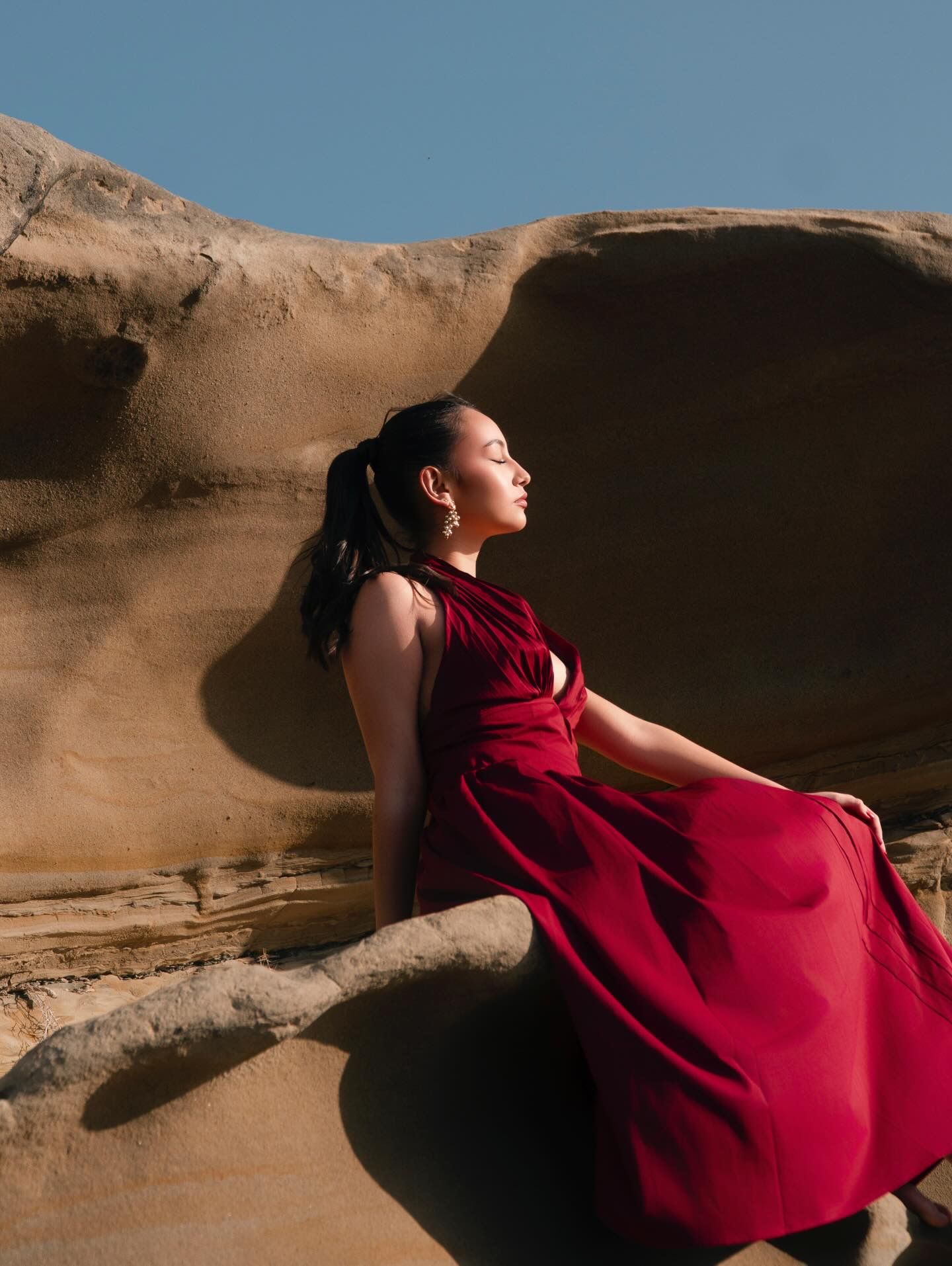 Woman in red dress poses gracefully against sandstone rock formations under a clear blue sky.