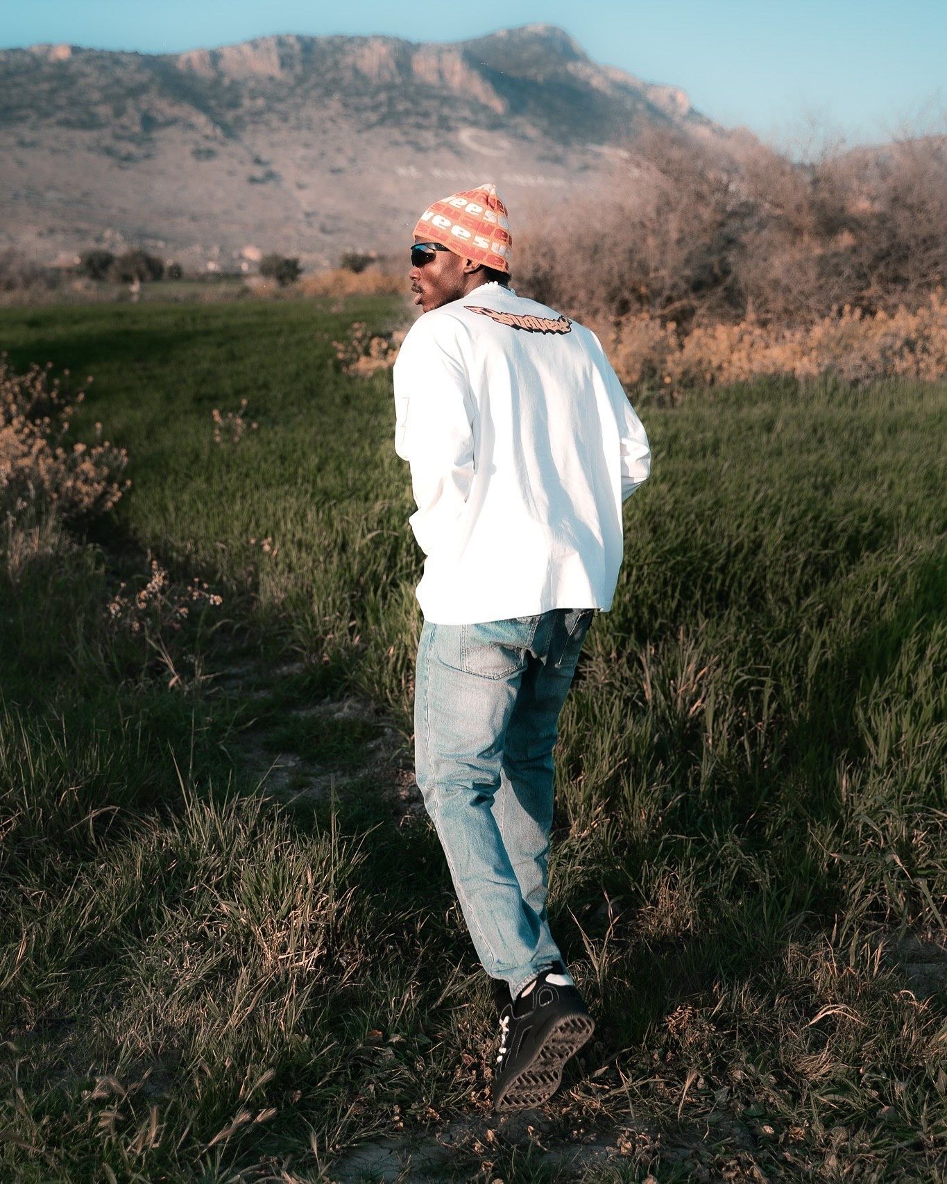 Man in white shirt, jeans, and orange beanie walks through green field with mountain backdrop.