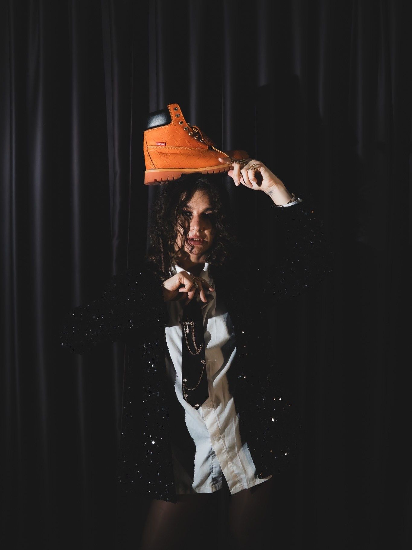 Woman holds orange boot above head wearing black sparkle jacket in dramatic dark studio editorial.