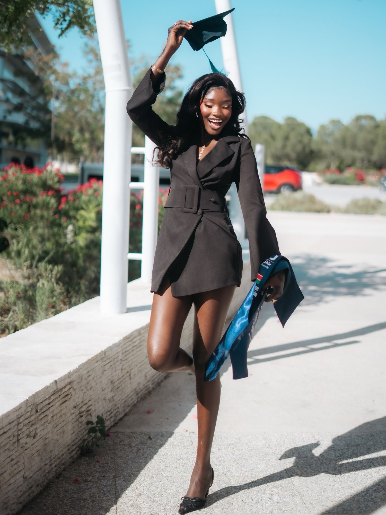 Smiling graduate in black blazer dress tosses cap in air while holding gown outdoors on sunny day.