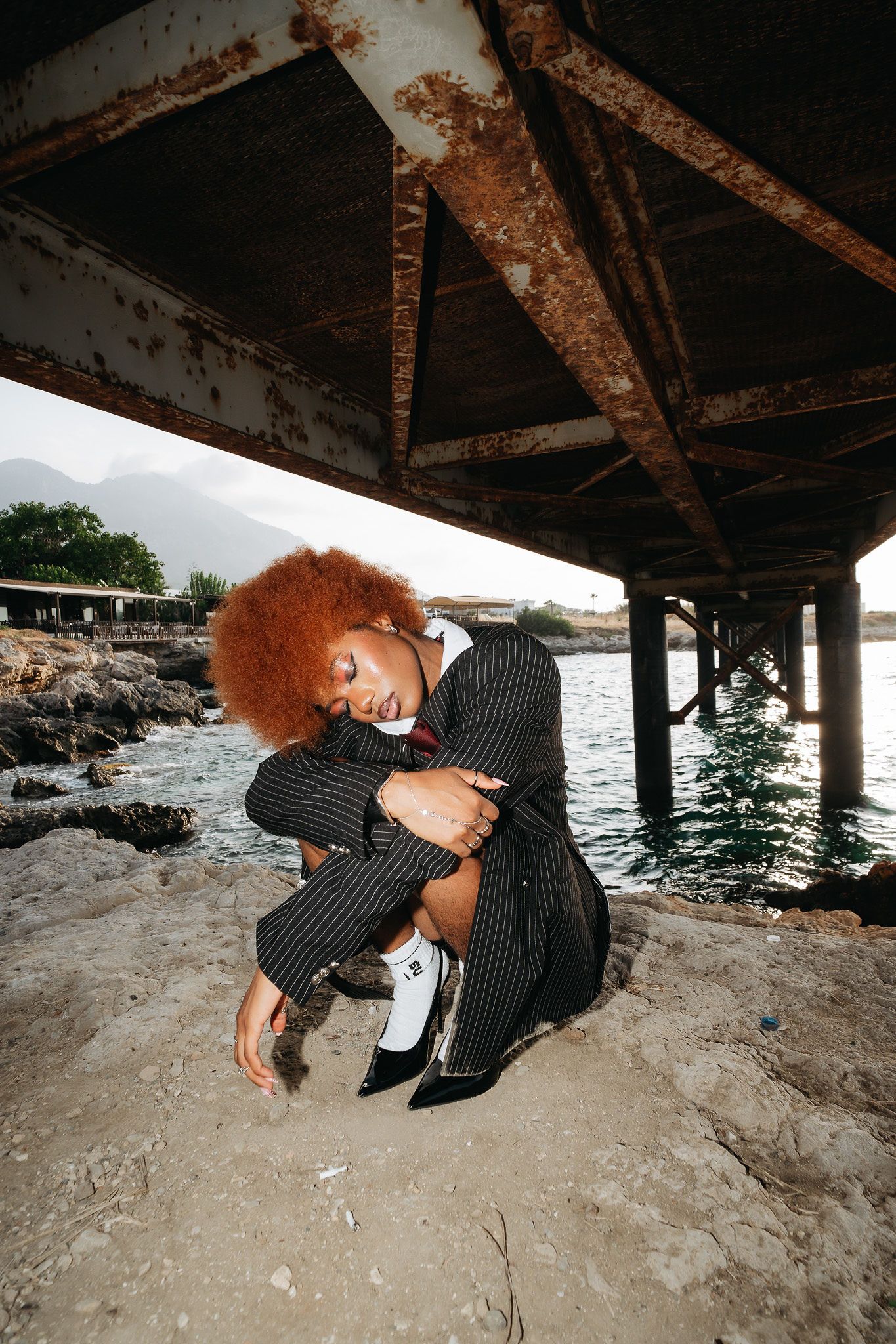 Woman with red afro in pinstripe suit crouching under a weathered pier near rocky shoreline.