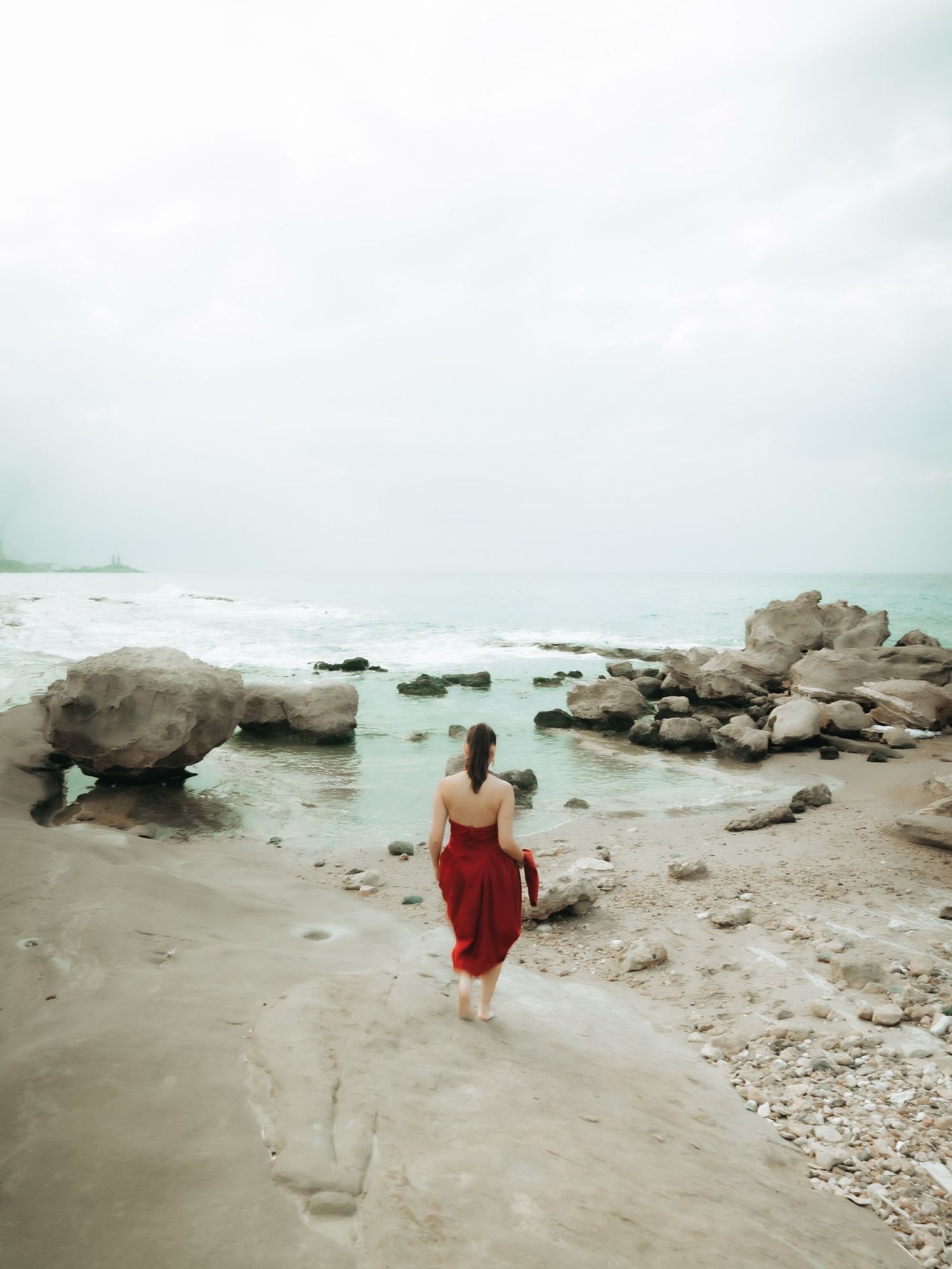 Woman in red dress walking toward rocky beach shoreline with turquoise waves under overcast sky.