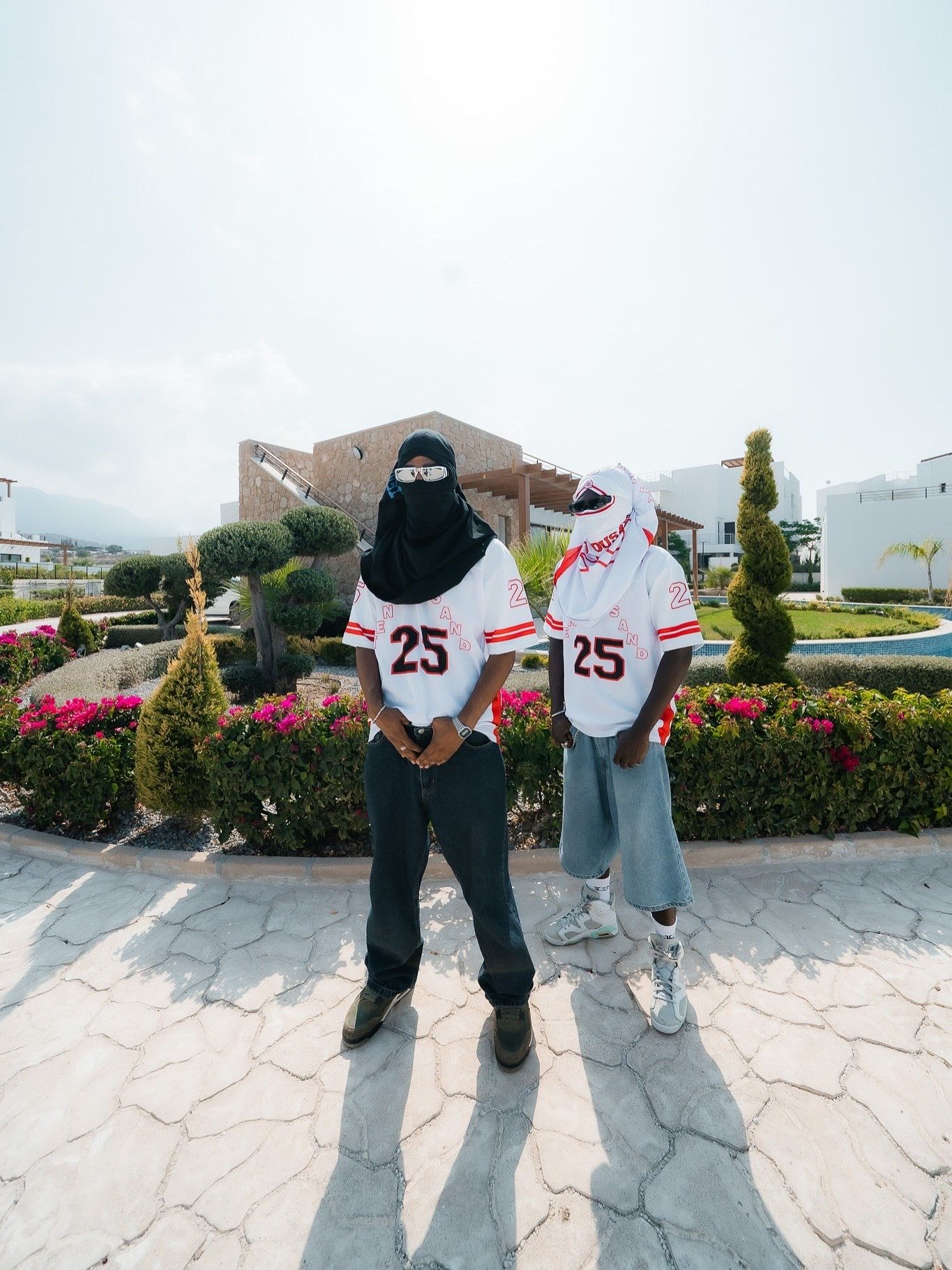 Two people wearing matching white baseball jerseys numbered 25 pose outdoors near a garden with colorful flowers.