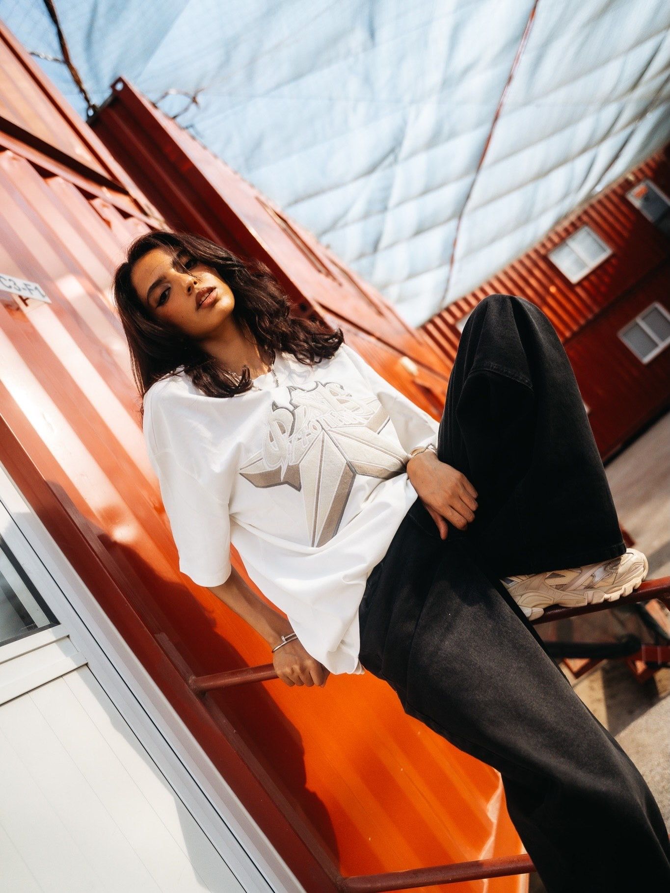 Woman in white top and black wide-leg pants sitting on orange shipping container in urban setting.