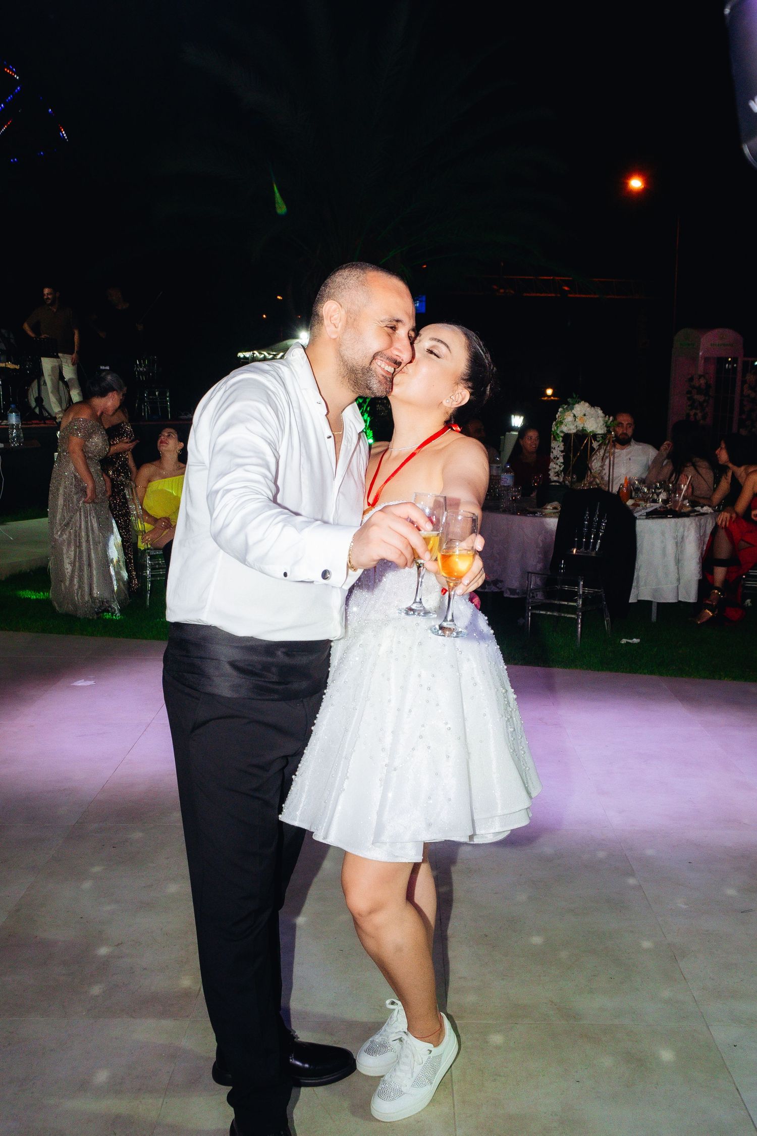 Couple sharing a romantic kiss on the dance floor at an outdoor evening wedding reception, holding champagne glasses.