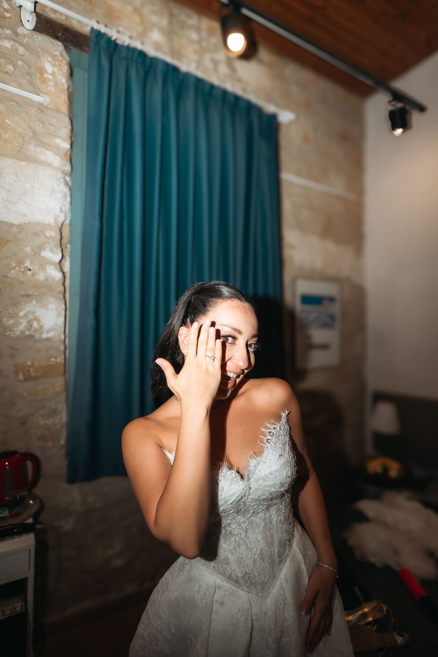 Emotional bride in lace wedding dress covering face with hand, standing in rustic stone room with teal curtains.
