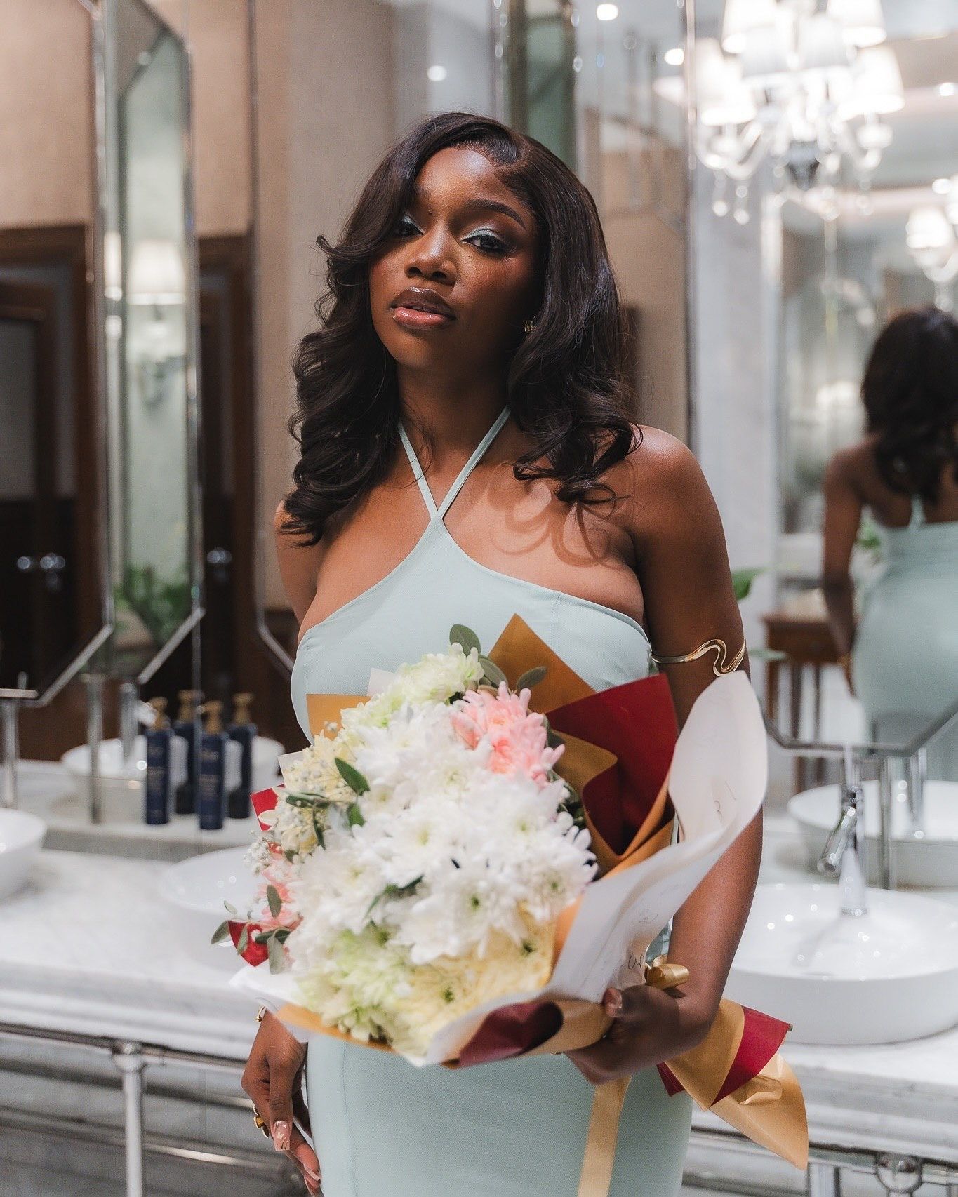 Elegant woman in mint halter dress holding a bouquet of white and pink flowers in a luxurious bathroom setting.