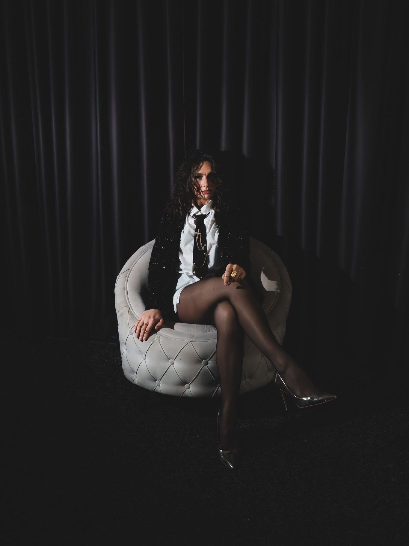 Woman in black blazer white shirt and silver heels sits crossed-legged in white tufted chair.