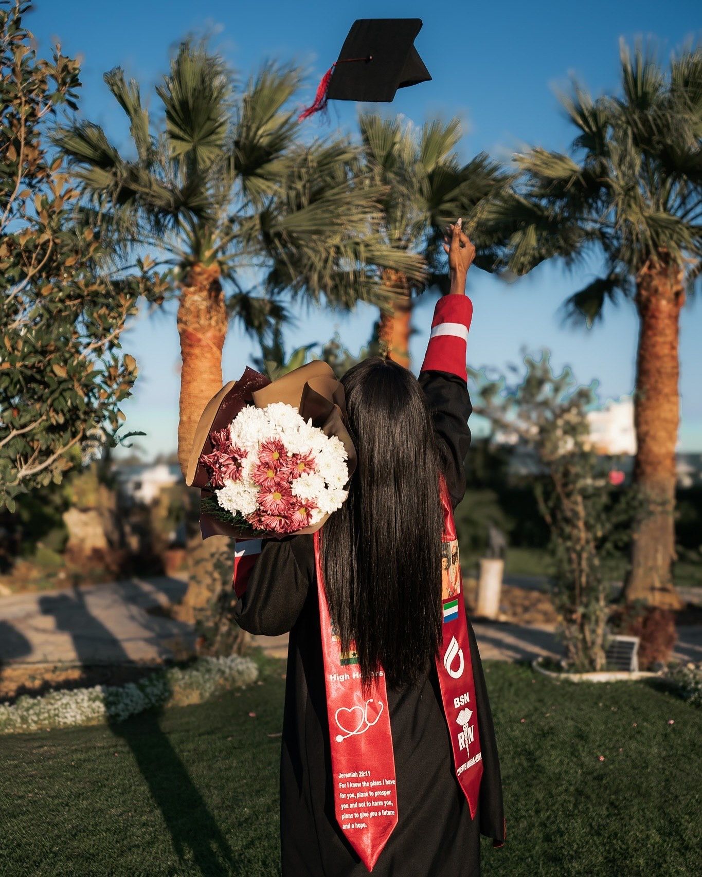 Graduate in black and red gown tosses mortarboard cap into air among palm trees.