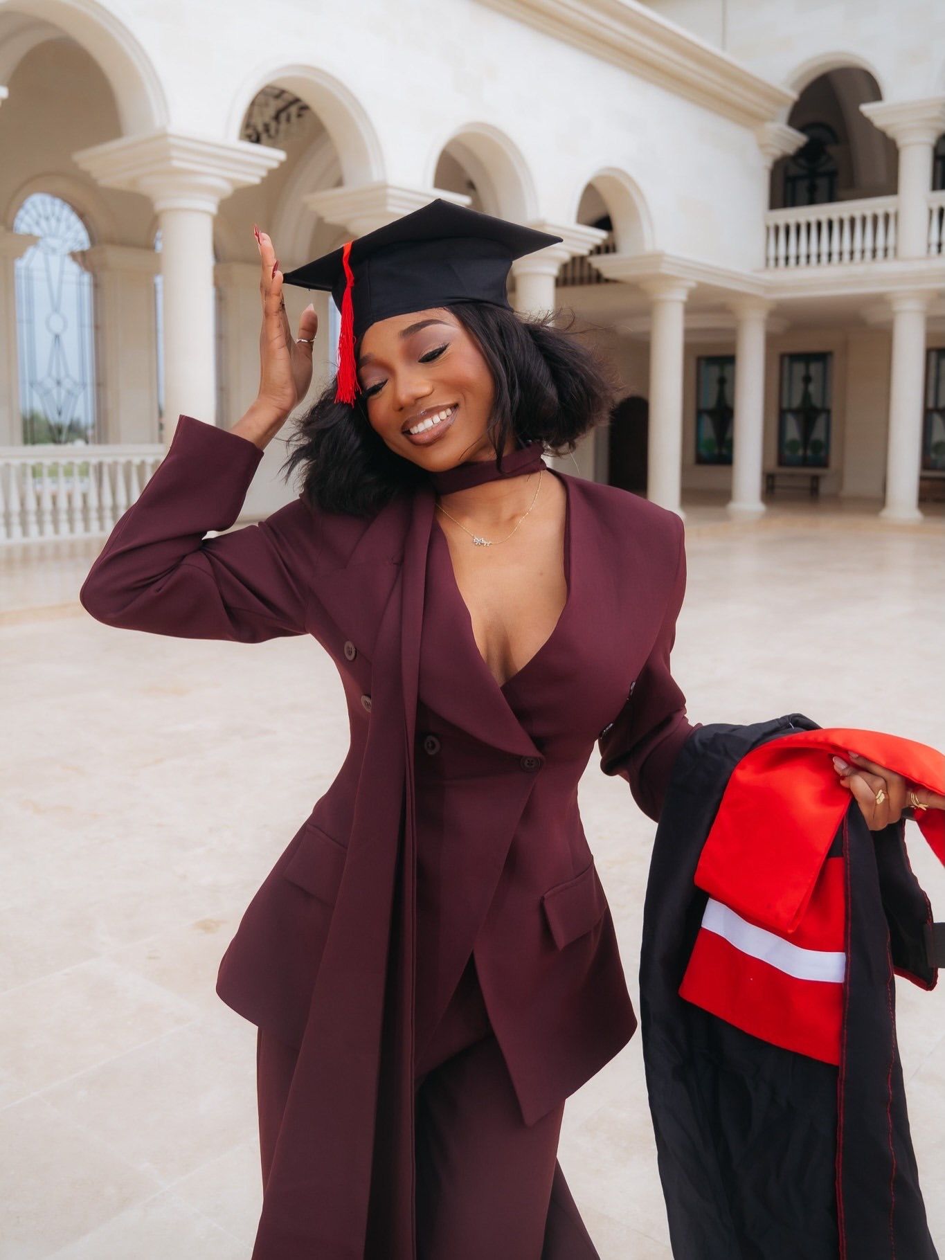 Joyful graduate in maroon pantsuit smiles while holding cap at elegant white columned building.