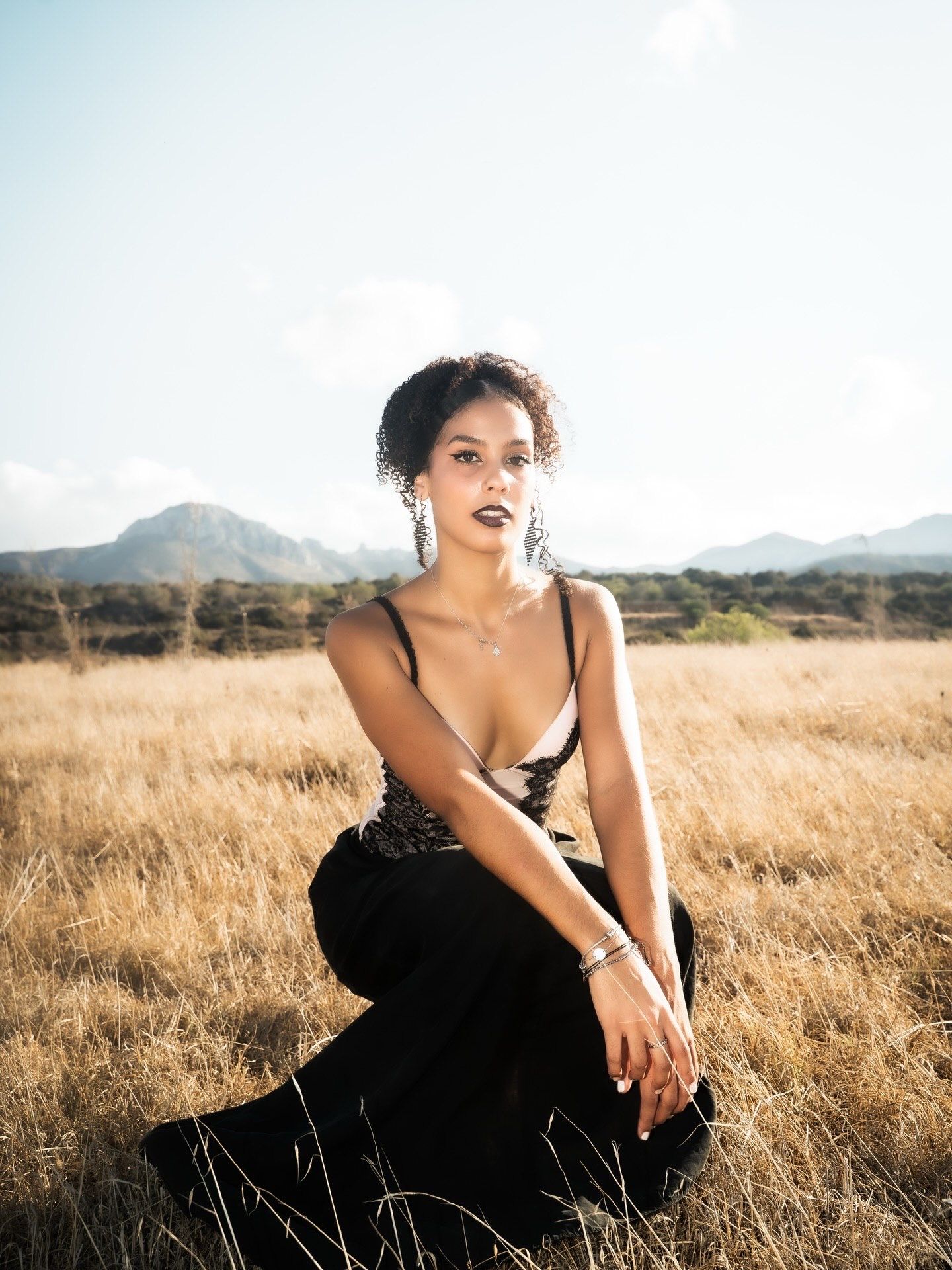 Woman in black dress sitting in golden field with mountains backdrop, wearing statement earrings and necklace.