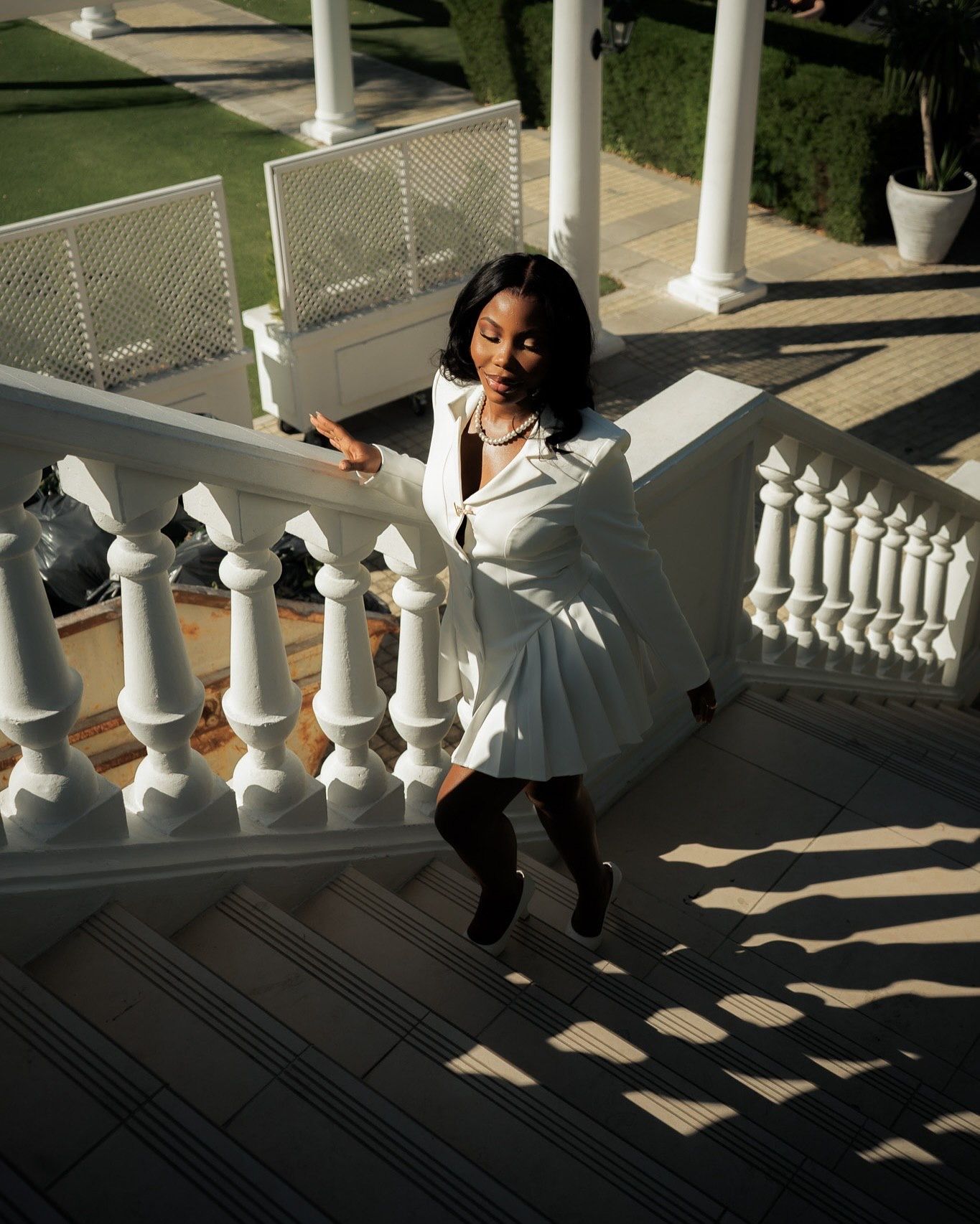 Woman in white coat descending elegant white marble staircase with ornate balusters at a luxury estate.