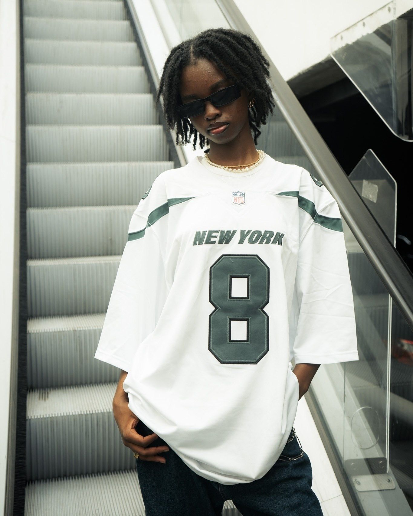 Woman wearing oversized New York Jets NFL jersey number 8 standing near escalator wearing sunglasses.
