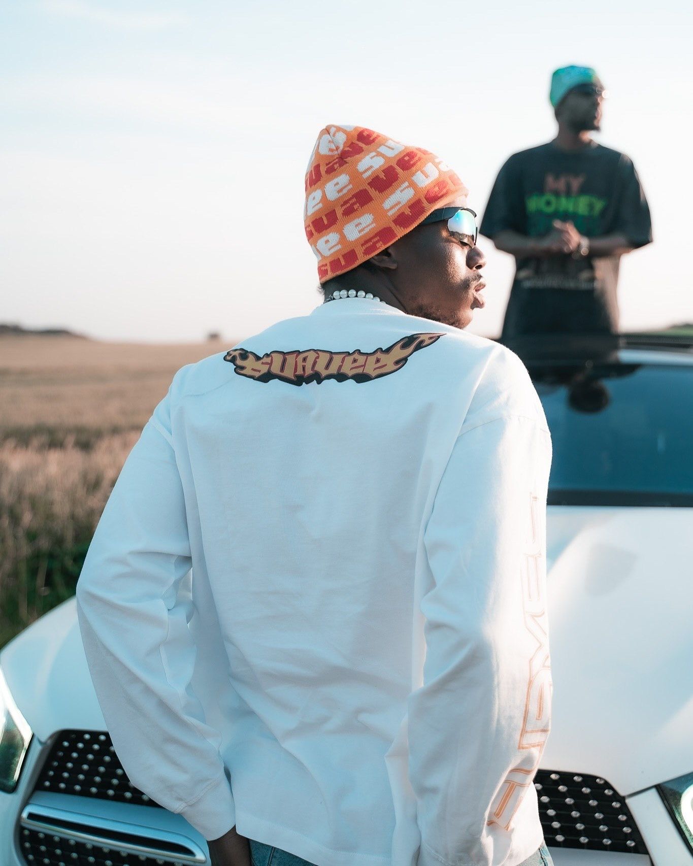 Two men near a white car outdoors, one wearing an orange beanie and white jacket with a graphic, another in a teal shirt.
