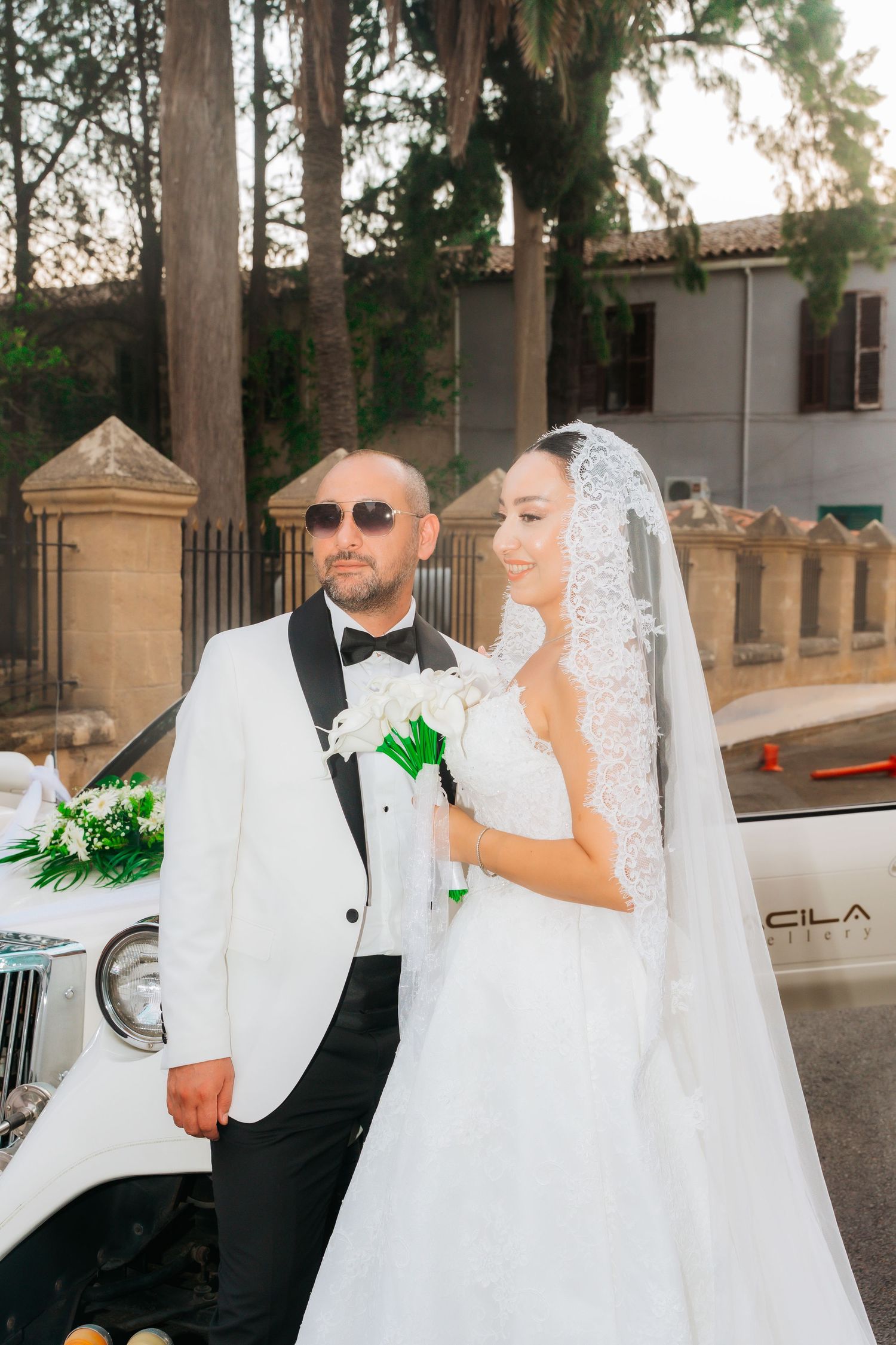 Bride in white gown and veil holds bouquet beside bald groom in white tuxedo near classic car outdoors.