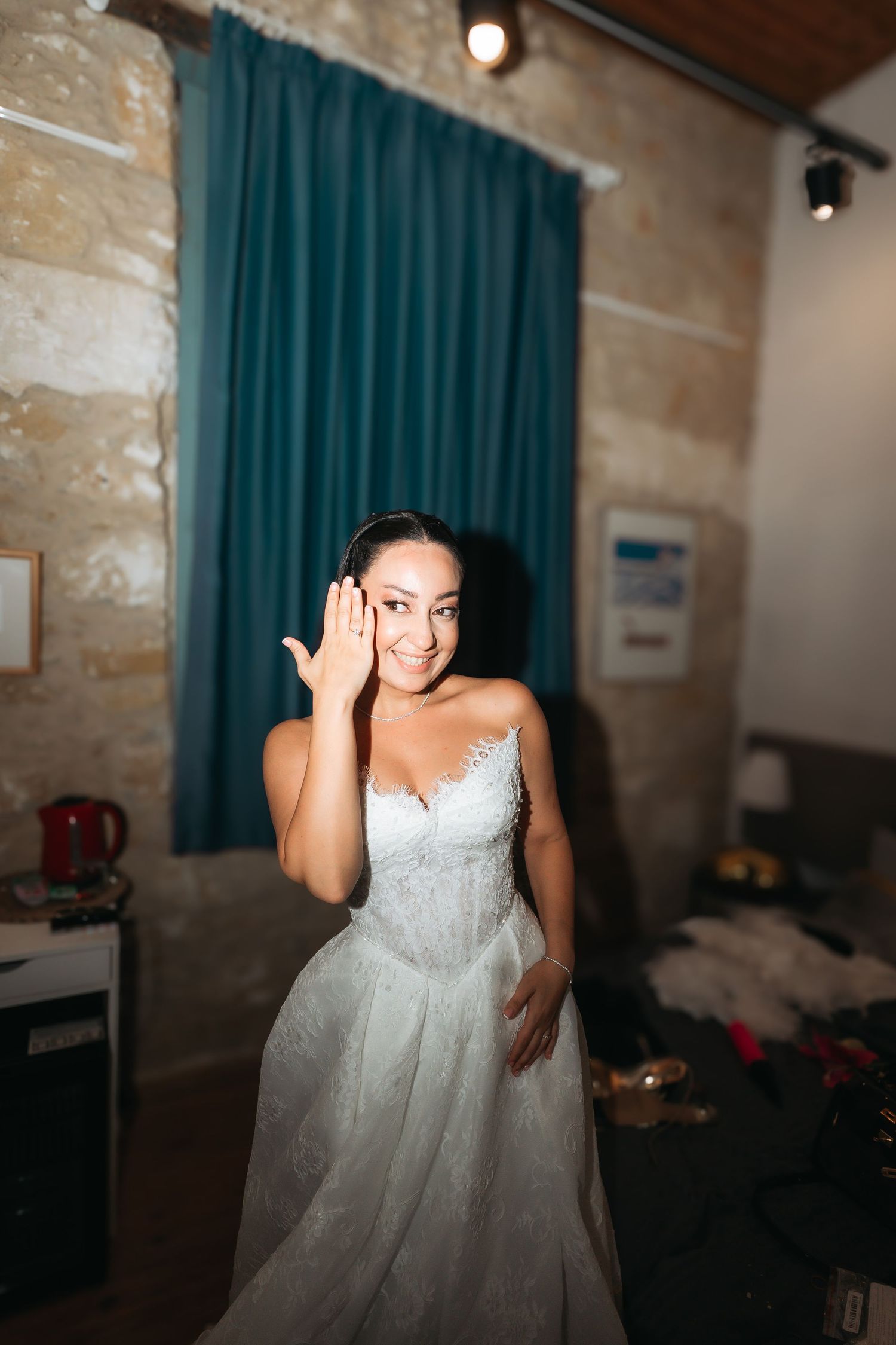 Smiling bride in white lace wedding dress adjusting her hair in a warmly lit room with teal curtains.