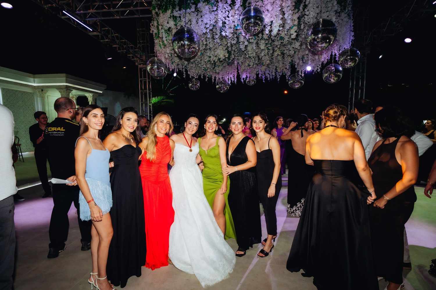Group of women in elegant gowns posing together at a wedding reception with floral chandelier decor.