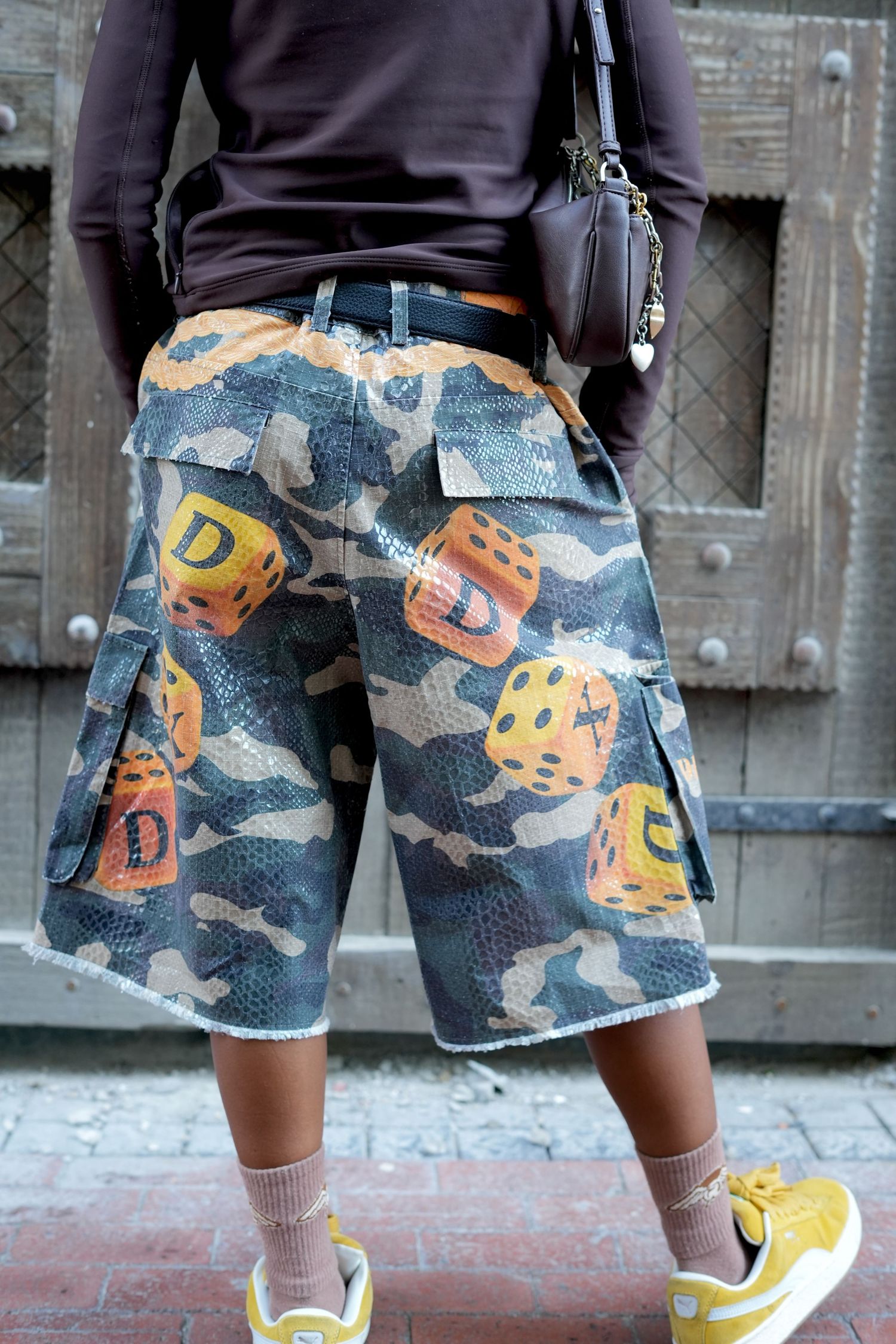 Person wearing camouflage cargo shorts with dice print, yellow sneakers, and a brown crossbody bag, standing outdoors.