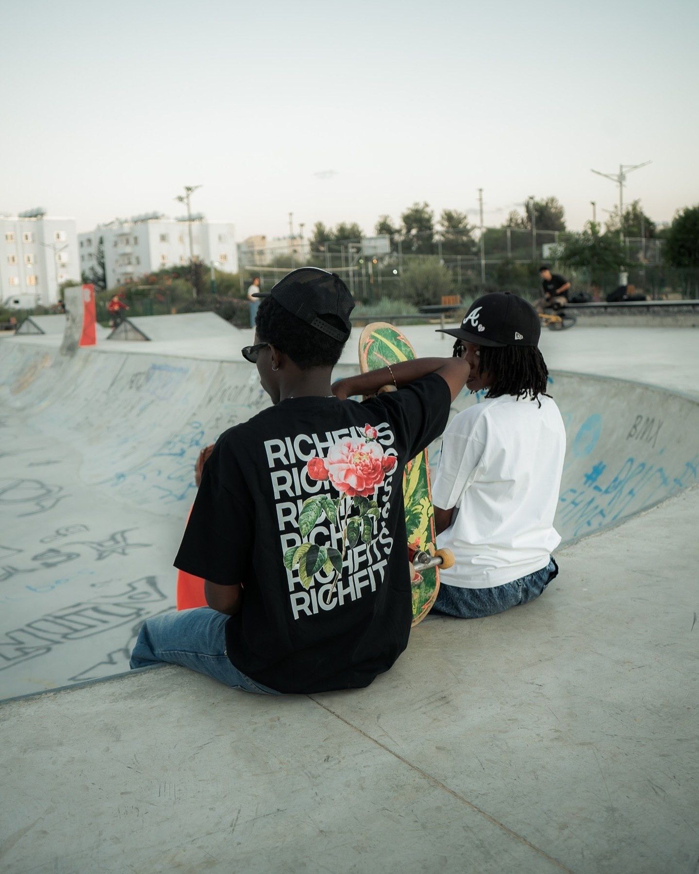 Two skaters wearing Riches caps sit at the edge of a concrete skate park bowl, one holding a colorful skateboard.