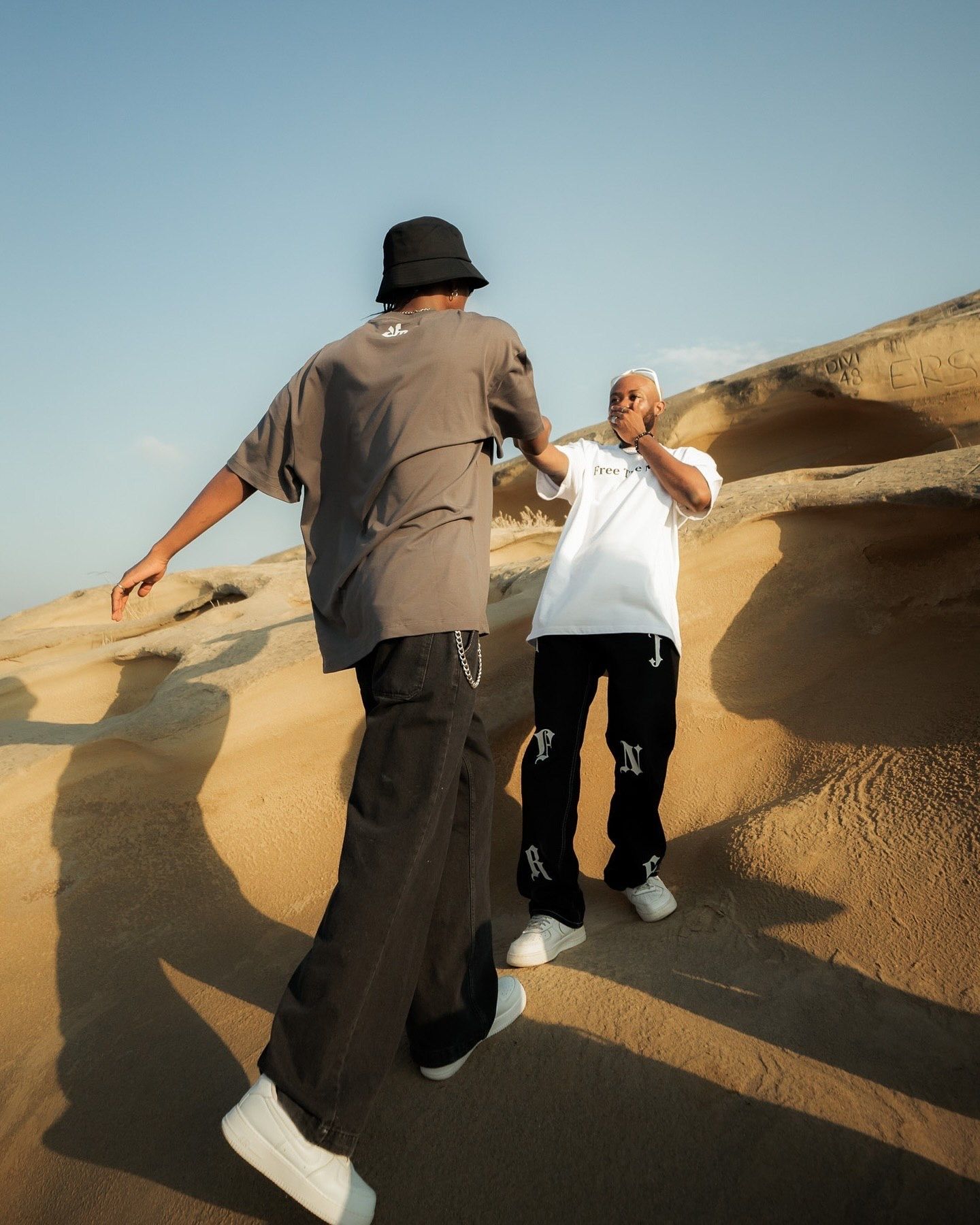 Two men in streetwear stand on sandy desert dunes, one wearing a bucket hat and gray tee, the other in a white tee and black pants.