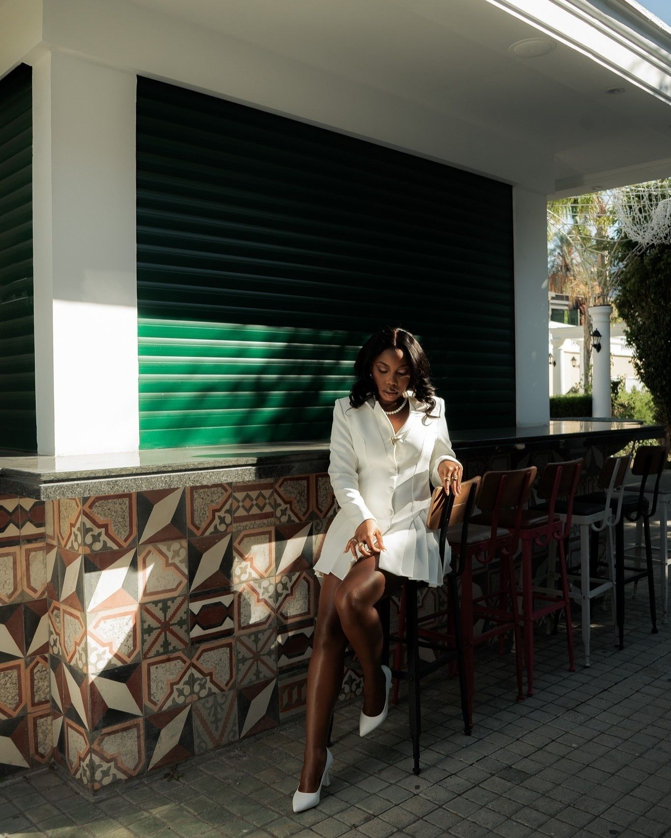 Woman in white blazer dress seated at a mosaic-tiled outdoor bar with green shutters backdrop.