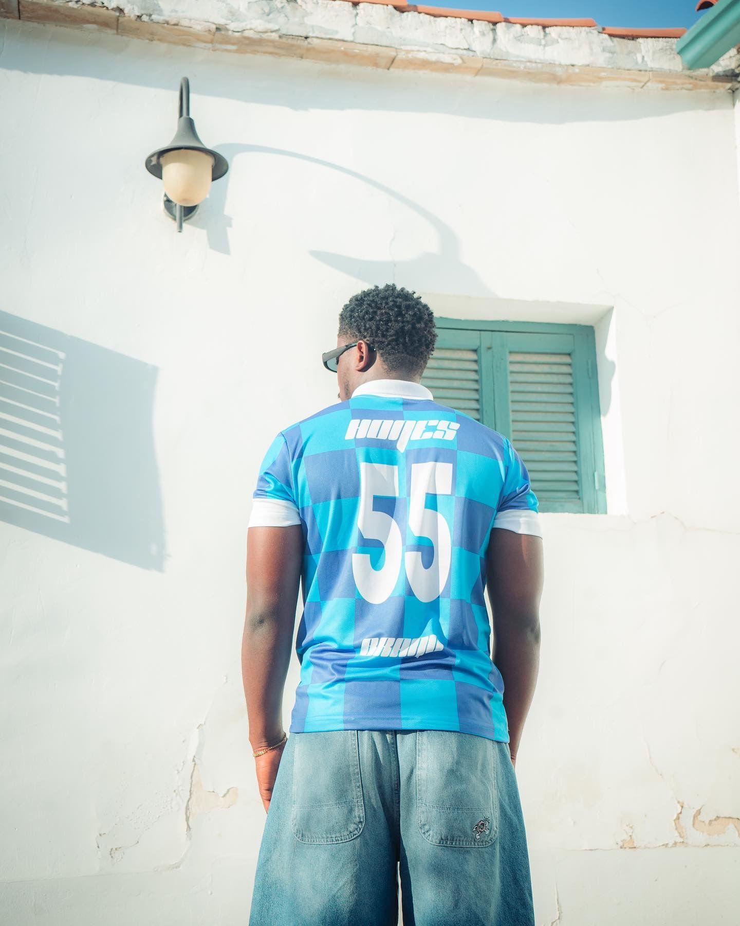 Young man in blue #55 jersey and jeans stands facing white wall with green shuttered window and wall lamp.